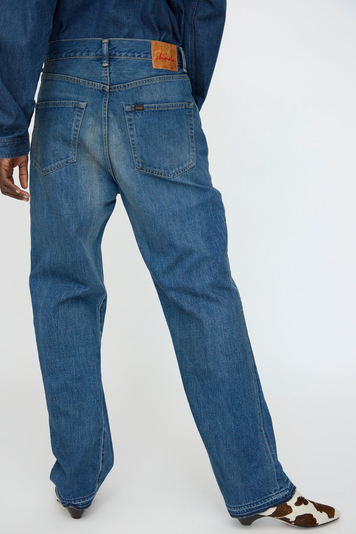 A person is seen from the back wearing Chimala's 13oz. Selvedge Denim Monroe Cut in Dark Distress jeans with a brown leather patch, styled with a denim jacket and cow-print shoes for a high-quality, fashion-forward look.