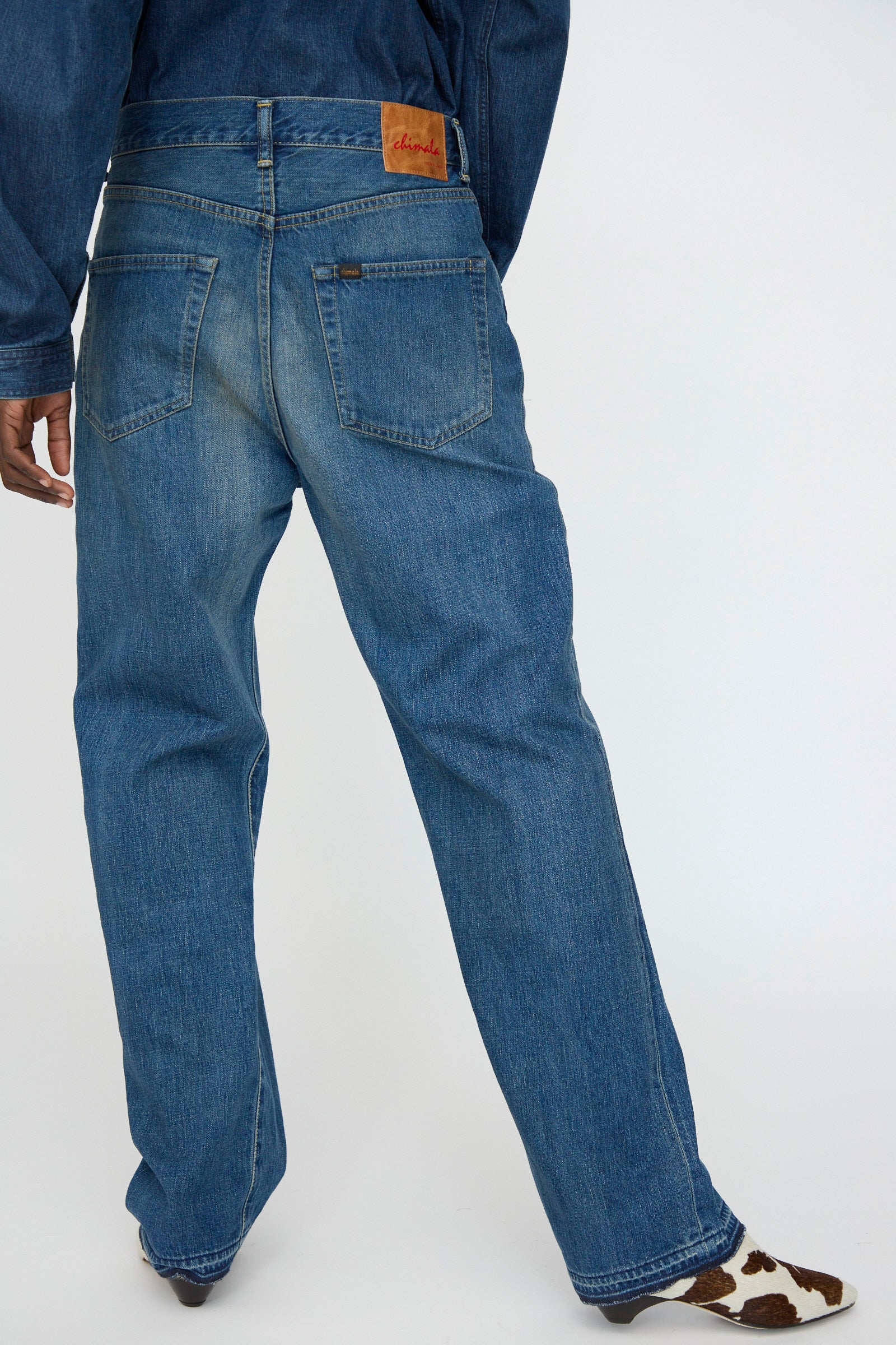 A person is seen from the back wearing Chimala's 13oz. Selvedge Denim Monroe Cut in Dark Distress jeans with a brown leather patch, styled with a denim jacket and cow-print shoes for a high-quality, fashion-forward look.