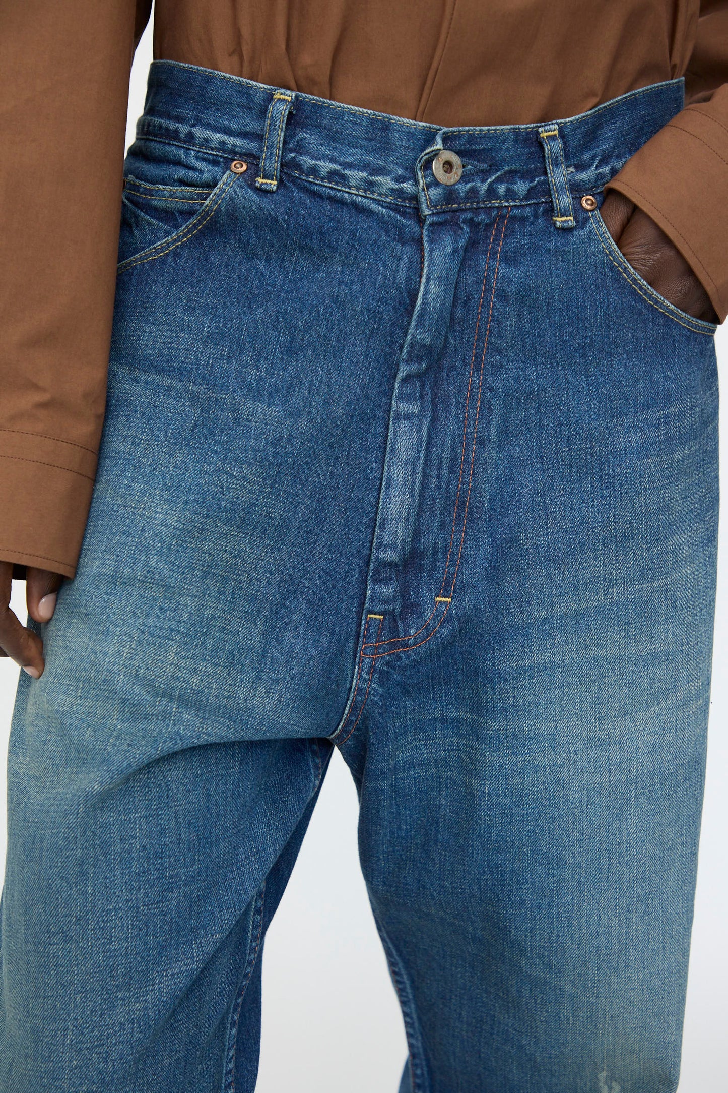 A person wears Chimala 13oz. Selvedge Denim Ranch Pants in Dark Wash—wide-leg, loose-fitting jeans with prominent seams—paired with a brown long-sleeve shirt, one hand in the pocket.
