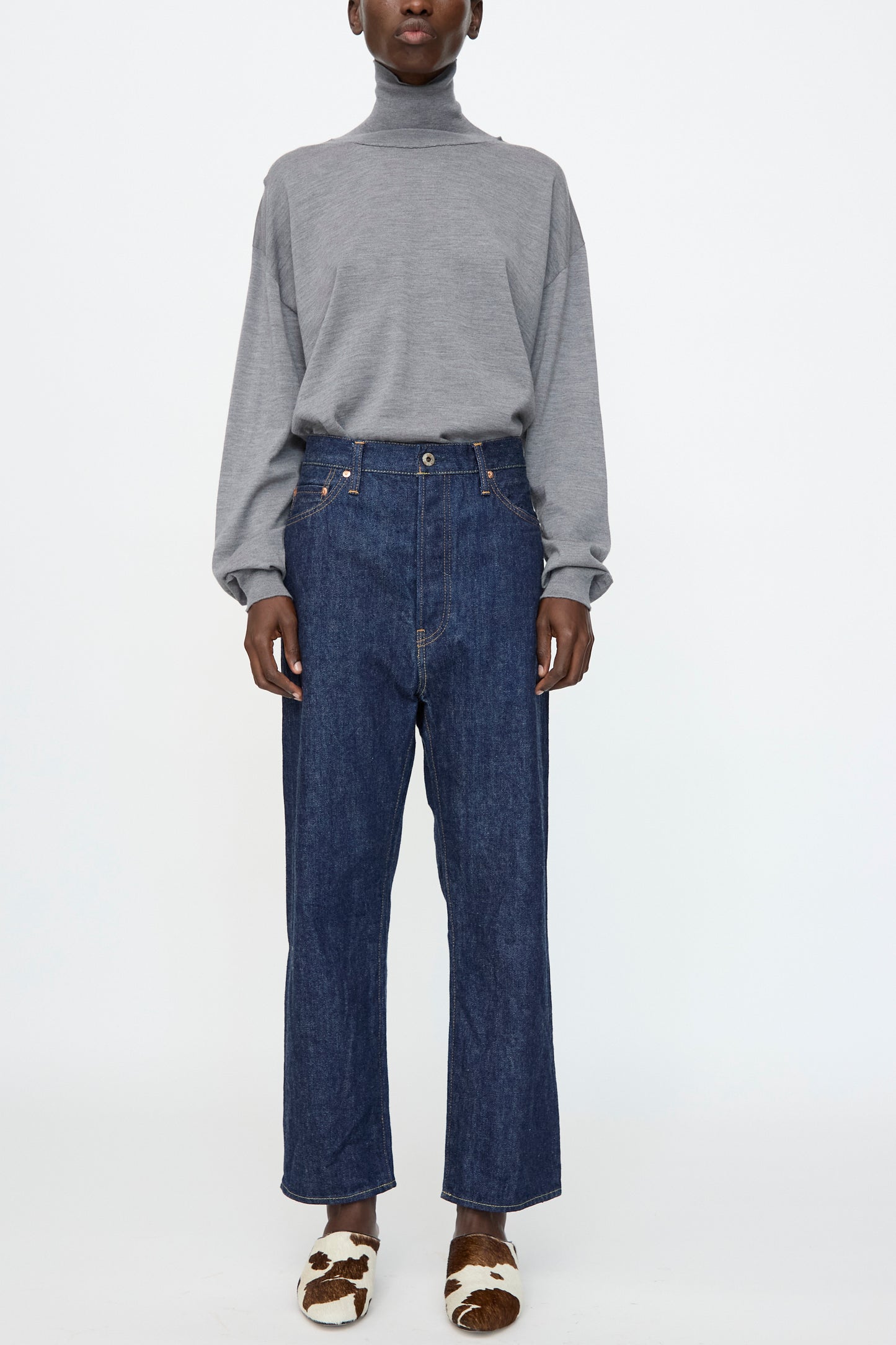 A person stands against a plain white background wearing a grey turtleneck, Chimala 13oz. Selvedge Denim Wide Tapered Cut in Rinse jeans, and brown-and-white cow print slippers.