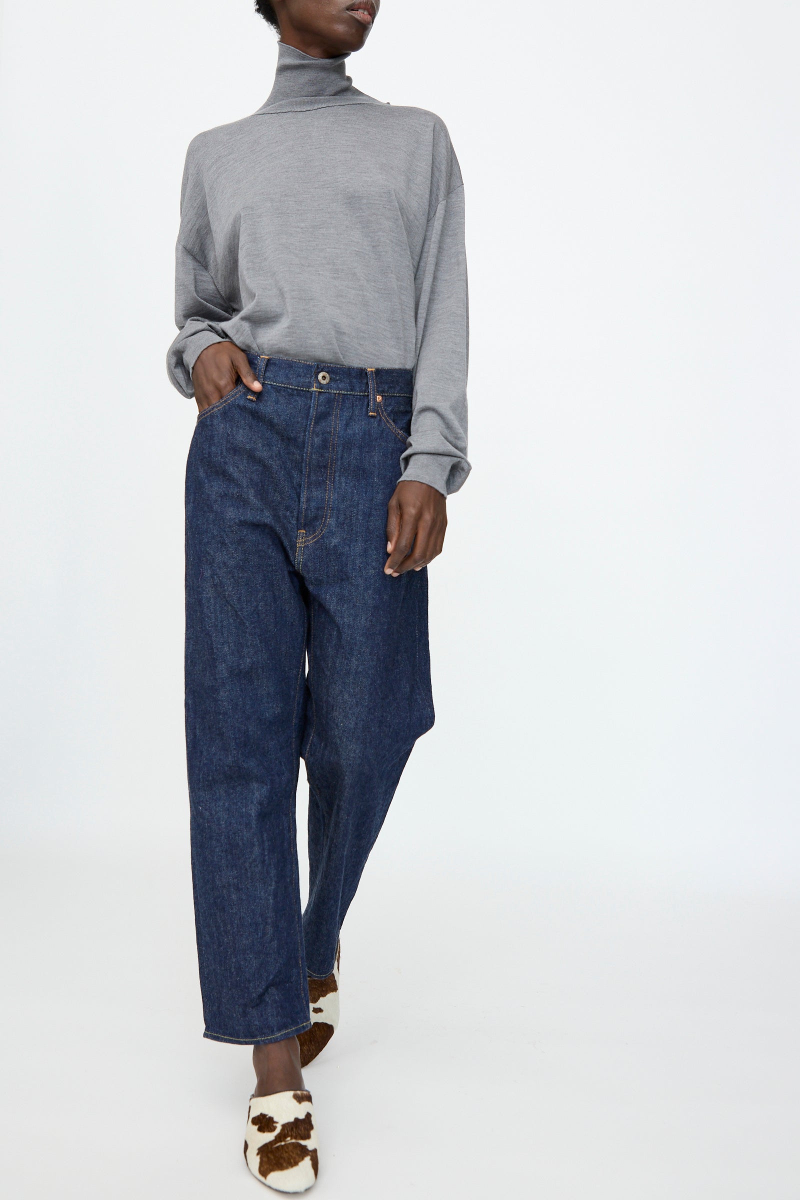 A person stands against a plain white background wearing a grey turtleneck, Chimala 13oz. Selvedge Denim Wide Tapered Cut in Rinse jeans, and brown-and-white patterned shoes.