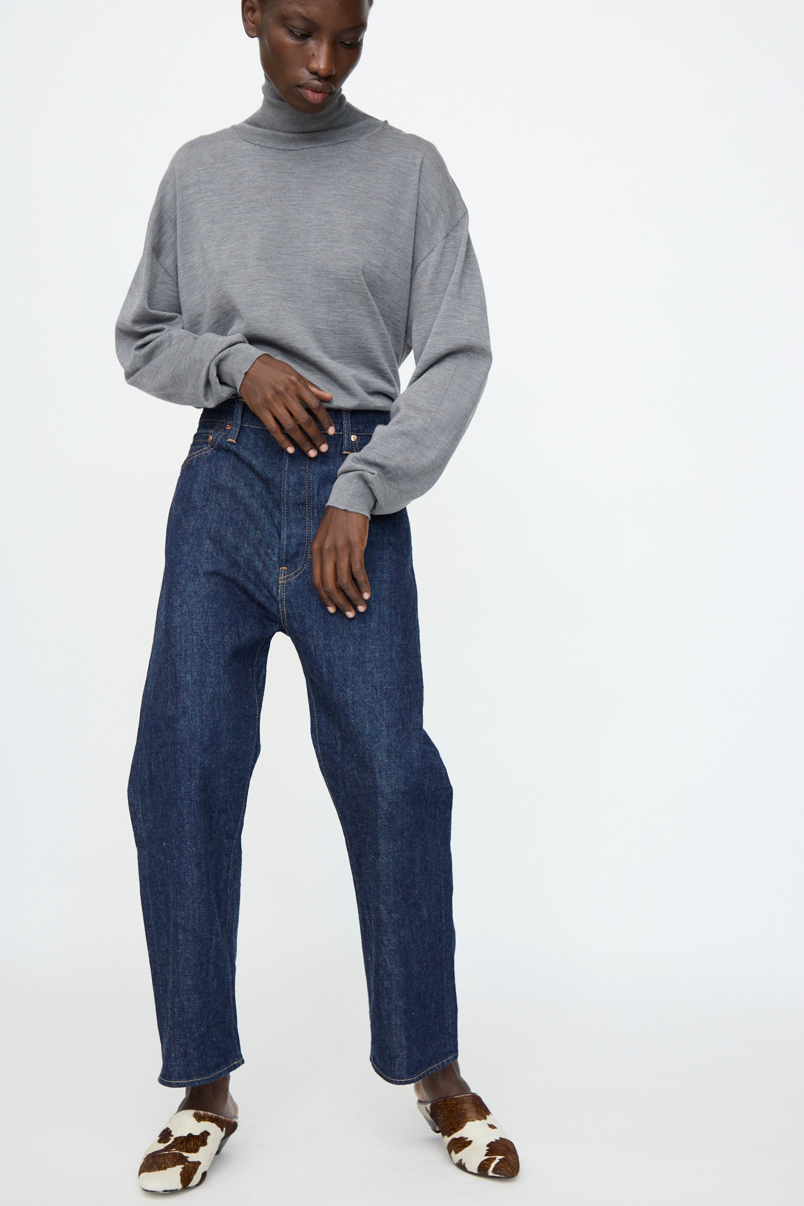 A person wearing a grey turtleneck, Chimala 13oz. Selvedge Denim Wide Tapered Cut in Rinse jeans, and brown-and-white cow print shoes stands against a plain white background.