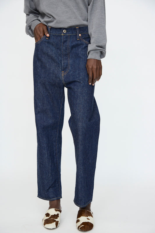 A person wearing a gray long-sleeve shirt, Chimala 13oz. Selvedge Denim Wide Tapered Cut in Rinse jeans, and brown and white cow print slippers.