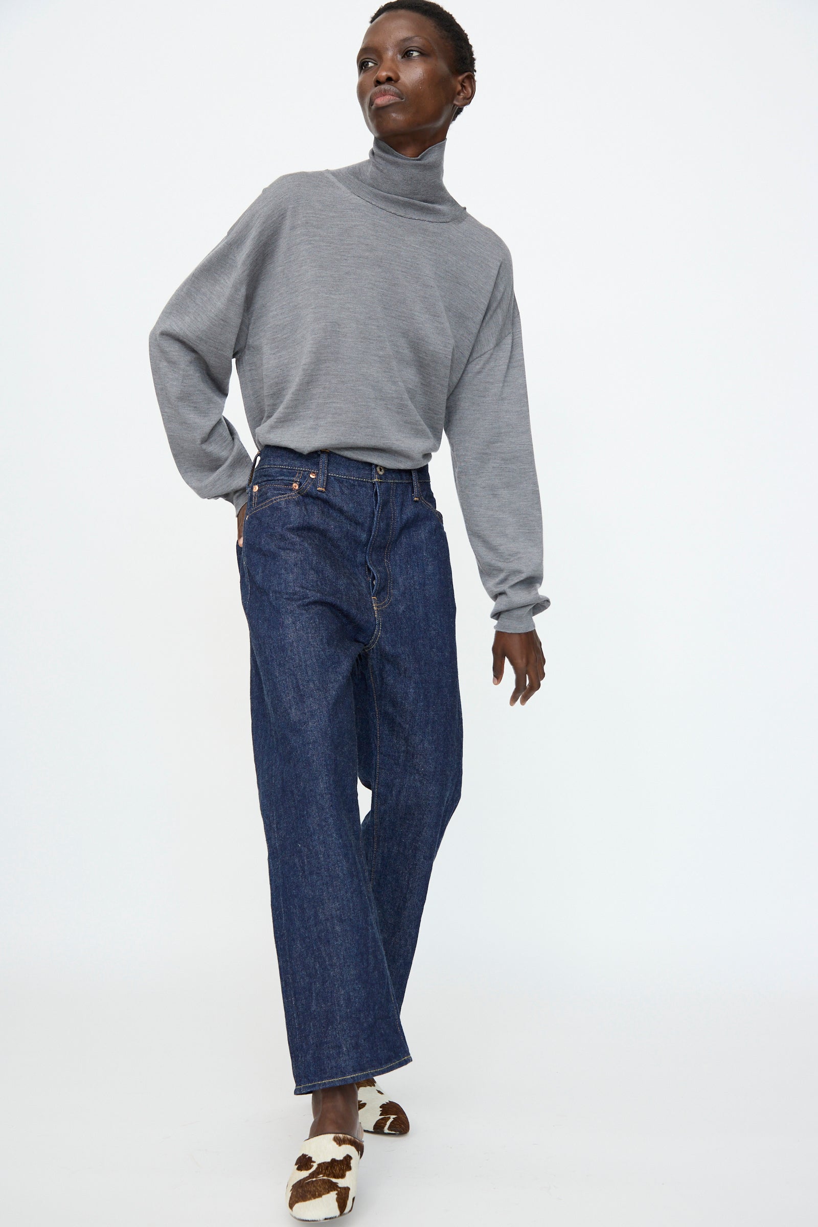 A person in a gray turtleneck sweater, Chimala 13oz. Selvedge Denim Wide Tapered Cut in Rinse jeans, and brown-and-white cow-print shoes stands against a plain white background.