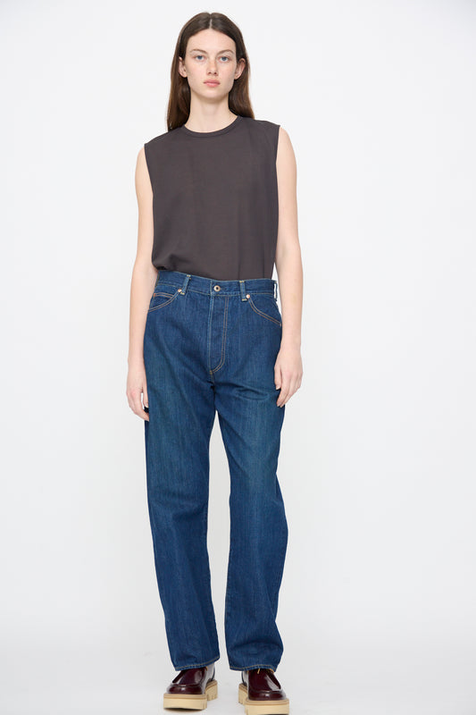 A person stands against a plain background, wearing a sleeveless black top, Chimala’s 13oz Selvedge Denim Monroe Cut in Dark Wash jeans, and burgundy shoes.