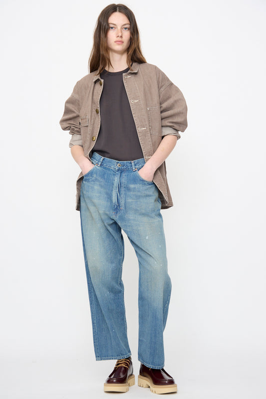 A person stands against a white background wearing a brown button-up jacket, black shirt, Chimala 13oz Selvedge Denim Ranch Pants in Work Light Blue, and brown platform shoes with hands in pockets.