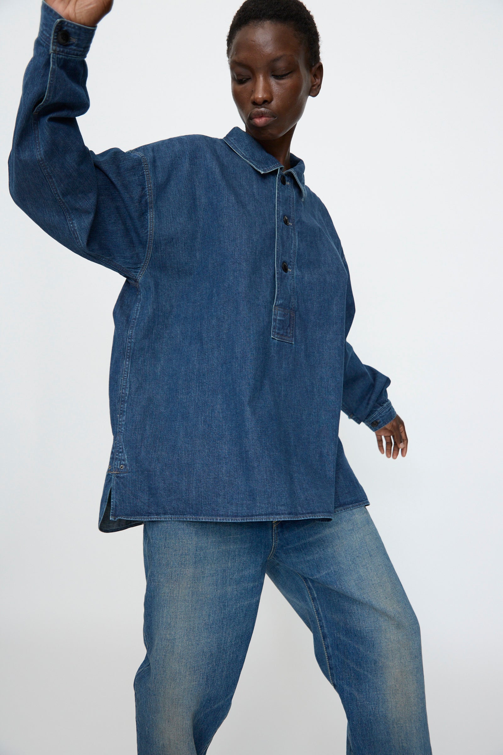 A person wearing the Chimala 8oz. Denim US Navydeck Shirt in Indigo with an oversized fit and jeans poses with one arm raised against a plain light background.
