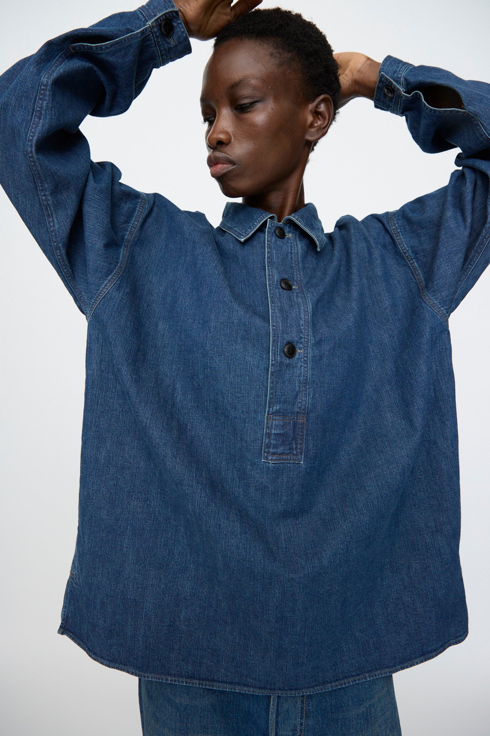 A person raises their arms against a plain background, wearing the Chimala 8oz. Denim US Navydeck Shirt in Indigo with black buttons and an oversized fit.