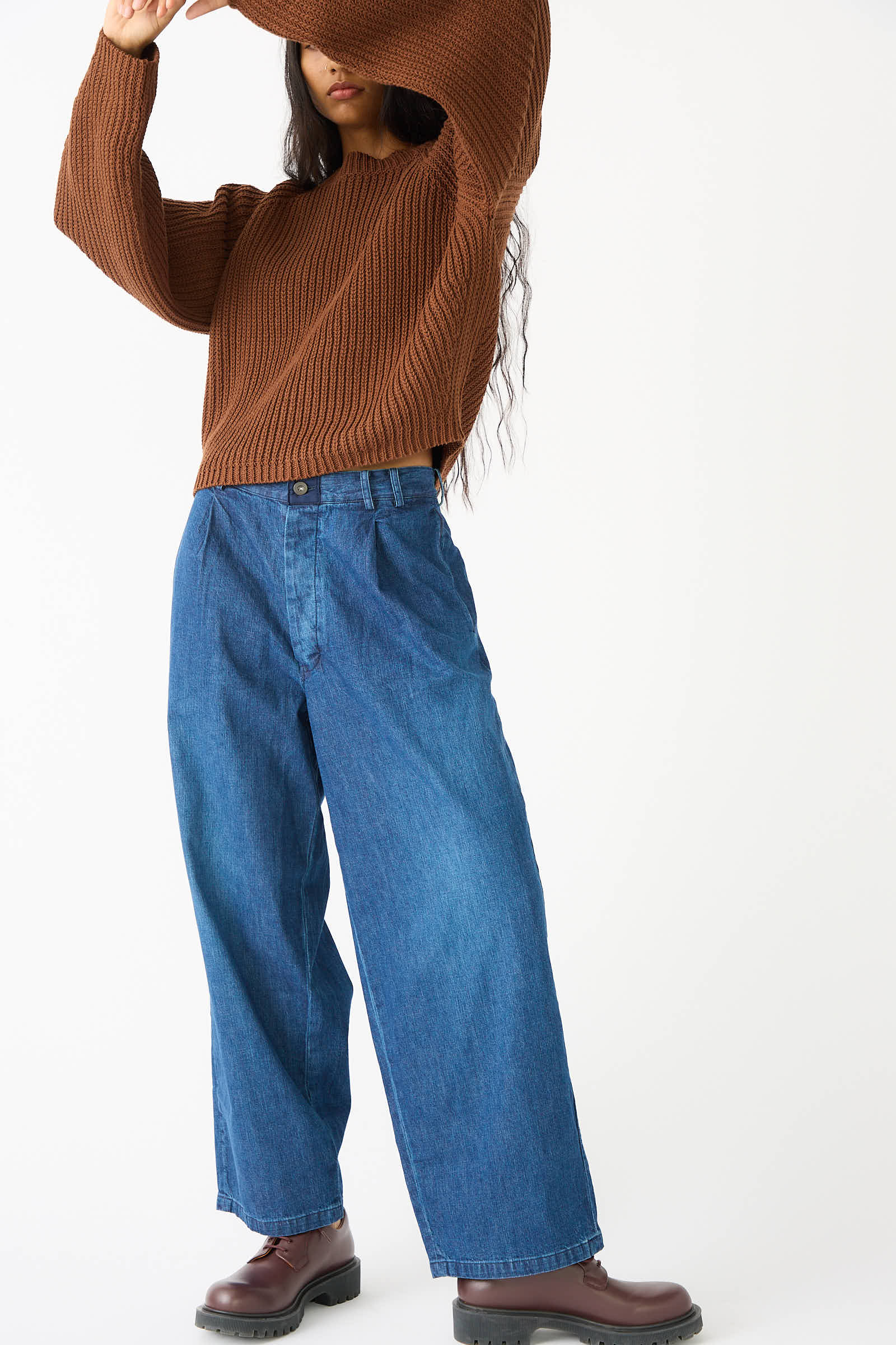 Person wearing a brown sweater, Chimala 8oz. Denim Trouser in Indigo, and brown shoes posing with arms raised, face partially covered by the sweater.