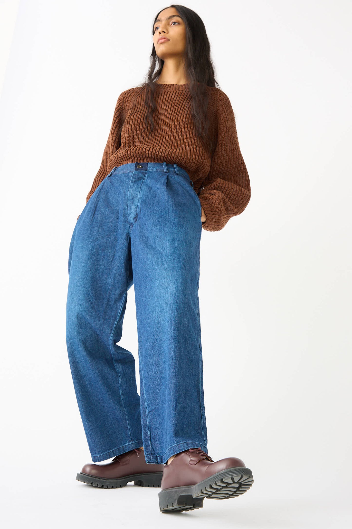 A person stands confidently wearing a brown sweater, the 8oz. Denim Trouser in Indigo by Chimala, and brown boots. They have long dark hair and look slightly upwards against a plain white background.