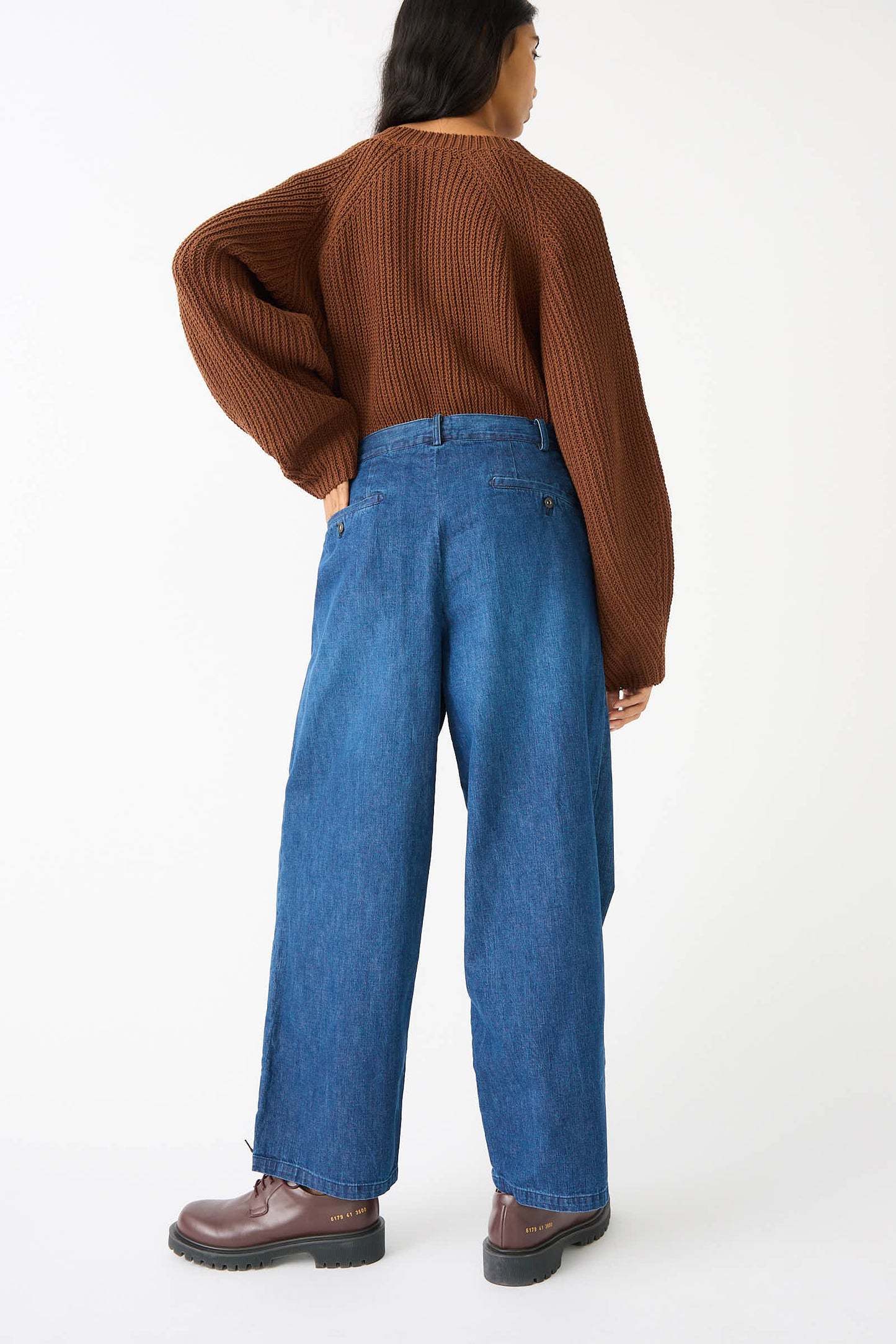 A person with long hair wearing a brown sweater, relaxed fit 8oz. Denim Trouser in Indigo by Chimala, and brown boots, standing and facing away from the camera against a white background.