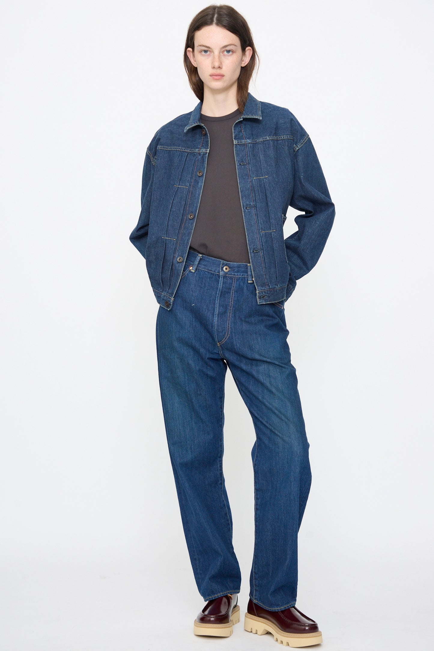 A person stands against a plain background, wearing the Chimala Amoskeag Denim Truck Jacket in Dark Wash, matching denim jeans, a dark t-shirt, and brown shoes with thick beige soles.