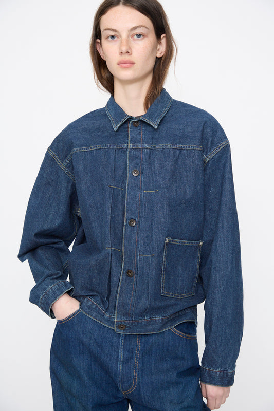 A person stands against a plain background wearing the Chimala Amoskeag Denim Truck Jacket in Dark Wash and matching pants, with one hand in their pocket.