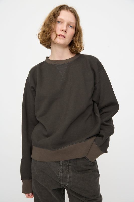 A person with wavy, shoulder-length hair is wearing the Chimala Brushed Fleece Crew Sweatshirt in Charcoal—a relaxed fit, long sleeve top crafted from brushed cotton fleece—paired with dark pants and standing against a plain light background with one hand in their pocket.
