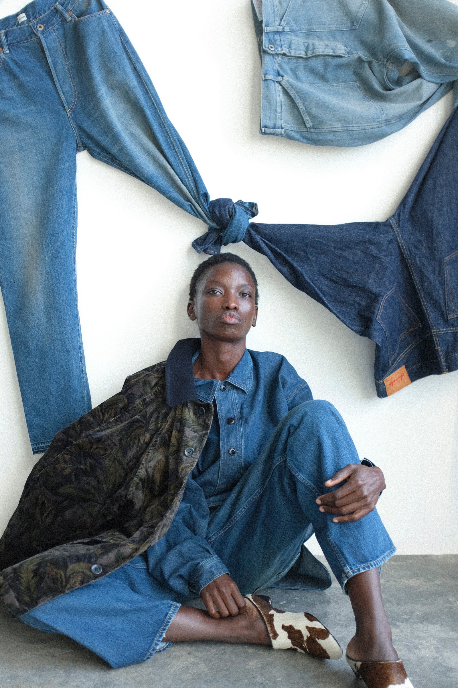 Person wearing a denim outfit with jeans and jacket, surrounded by denim clothing on a white wall.