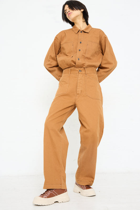 Woman wearing a Chimala Classic Drill US Army Work Trouser in Camel jumpsuit made from Japanese cotton canvas with chunky white and brown shoes, posing with her hands in pockets against a white background.