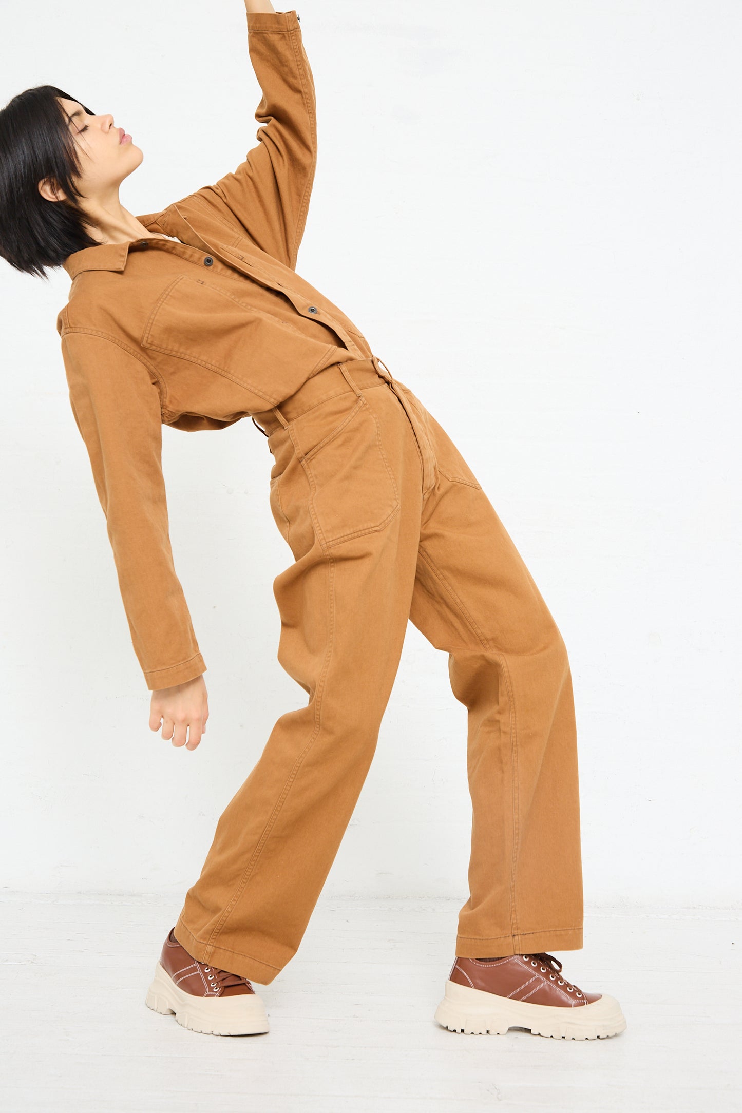 Woman in a Chimala Classic Drill US Army Work Trouser in Camel jumpsuit made from Japanese cotton canvas posing dynamically against a white background.