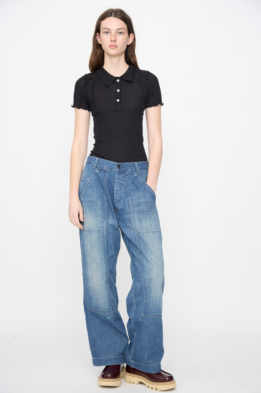 A person stands against a plain background wearing a black short-sleeve polo shirt, Chimala Denim Double Knee Painter Pants in Splushed Medium, and brown platform shoes with light soles.