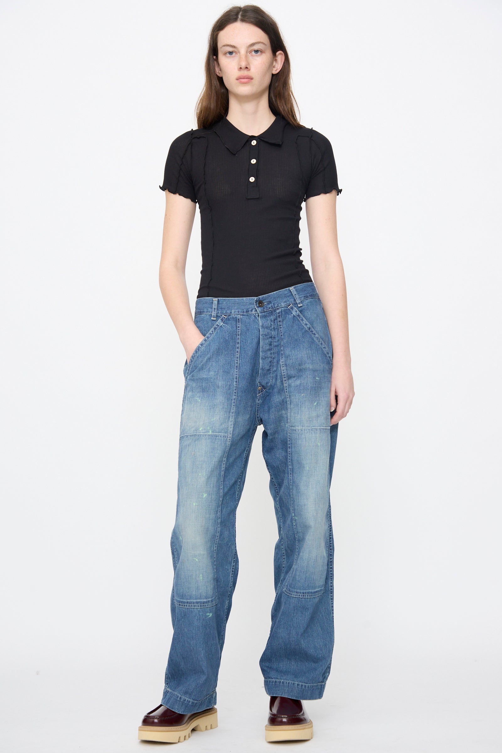 A person stands against a white background wearing a black short-sleeve polo, Chimala Denim Double Knee Painter Pants in Splushed Medium blue, and burgundy shoes. Their hands are in their pockets.