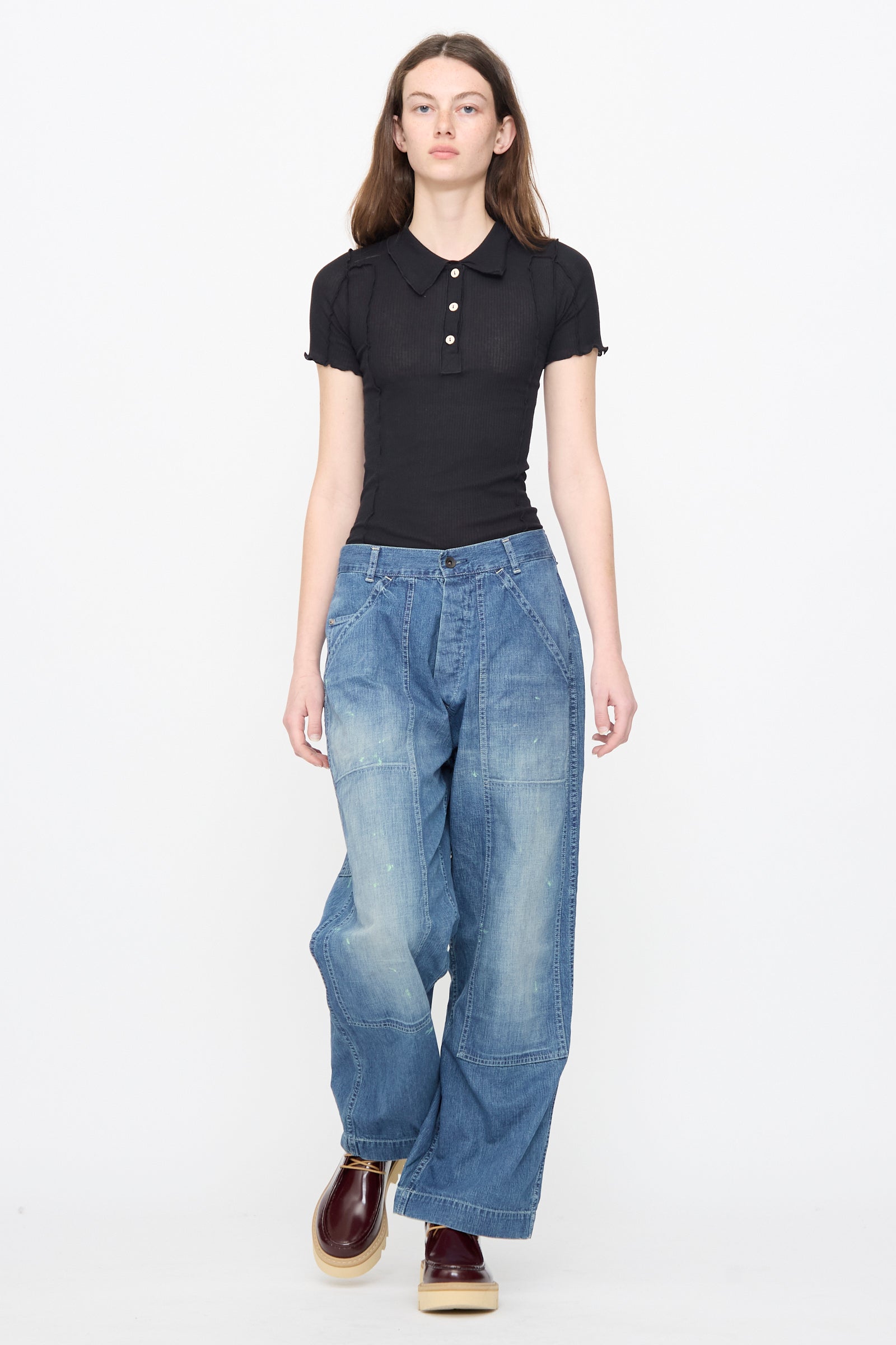 A person wears a fitted black short-sleeve polo, Chimala Denim Double Knee Painter Pants in Splushed Medium, and brown shoes, standing against a plain white background.