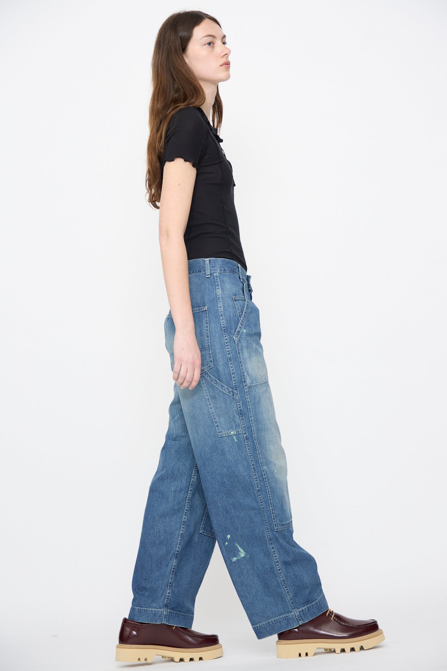 A woman stands sideways in a black short-sleeve top, Chimala Denim Double Knee Painter Pants in Splushed Medium, and brown platform shoes against a plain white background.