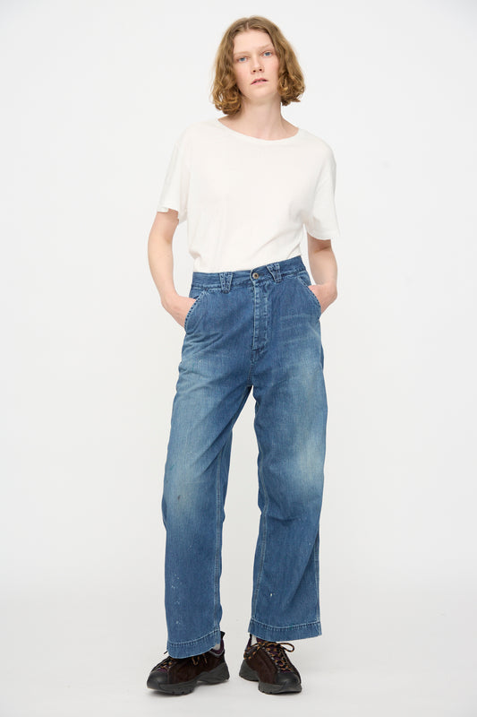 Person standing against a plain background, wearing a white t-shirt, Chimala’s Denim Painter Pant in Medium Engineer (a relaxed fit Japanese denim wide leg blue jean), and black shoes, with hands in pockets.