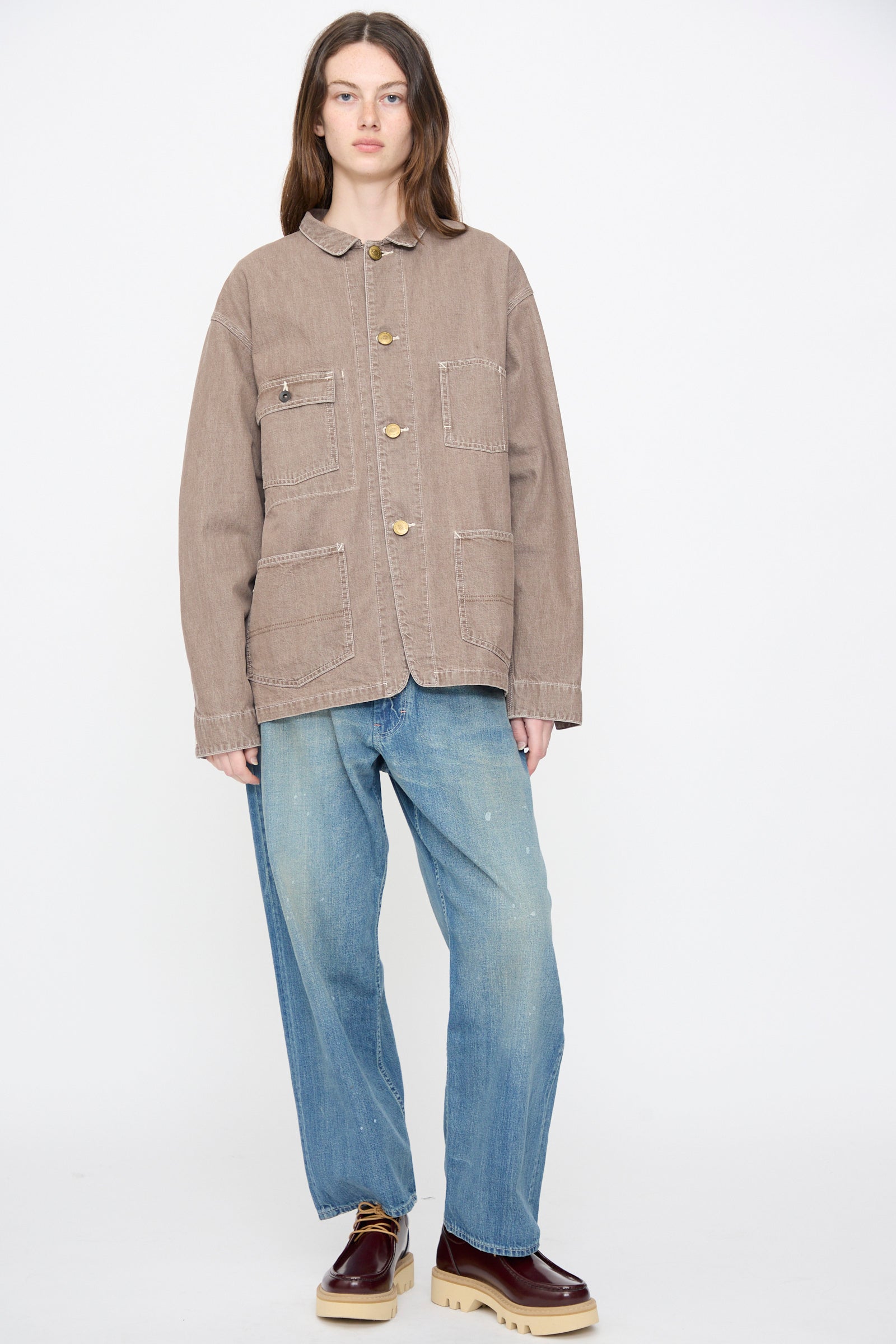 A person stands against a plain background wearing Chimala’s Denim Short Chore Jacket in Brown, styled with loose blue jeans and maroon platform shoes for an effortless, relaxed 100% cotton look.