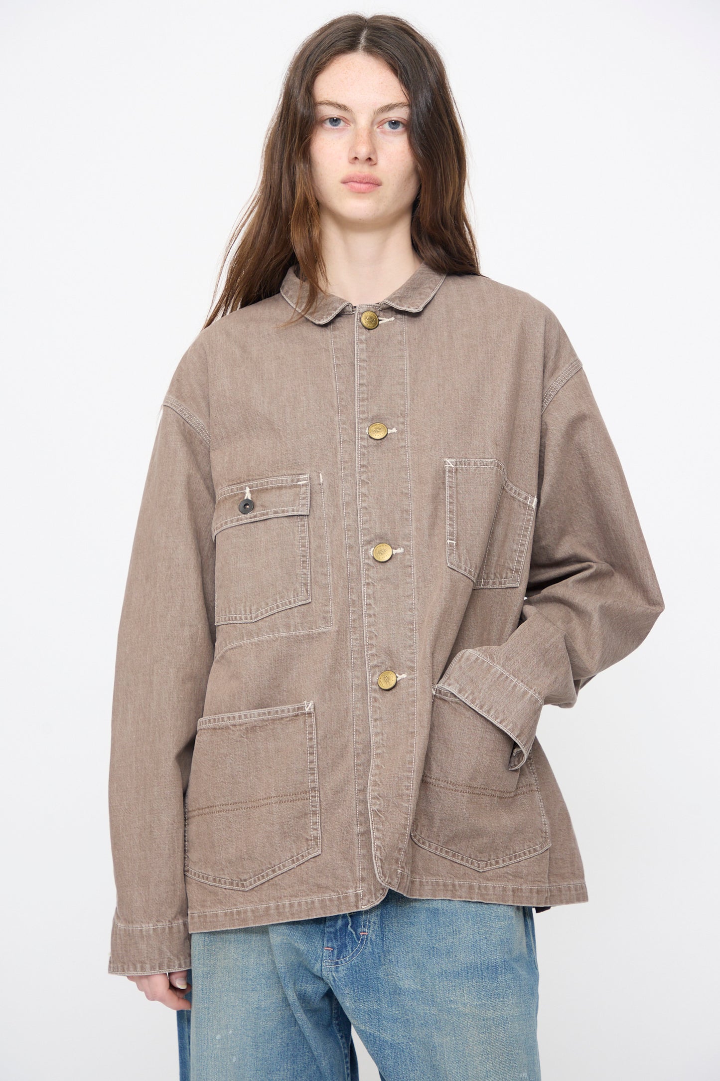 A person wears Chimala’s Denim Short Chore Jacket in Brown—a relaxed-fit, 100% cotton jacket with multiple front pockets—paired with blue jeans, standing against a plain white background.