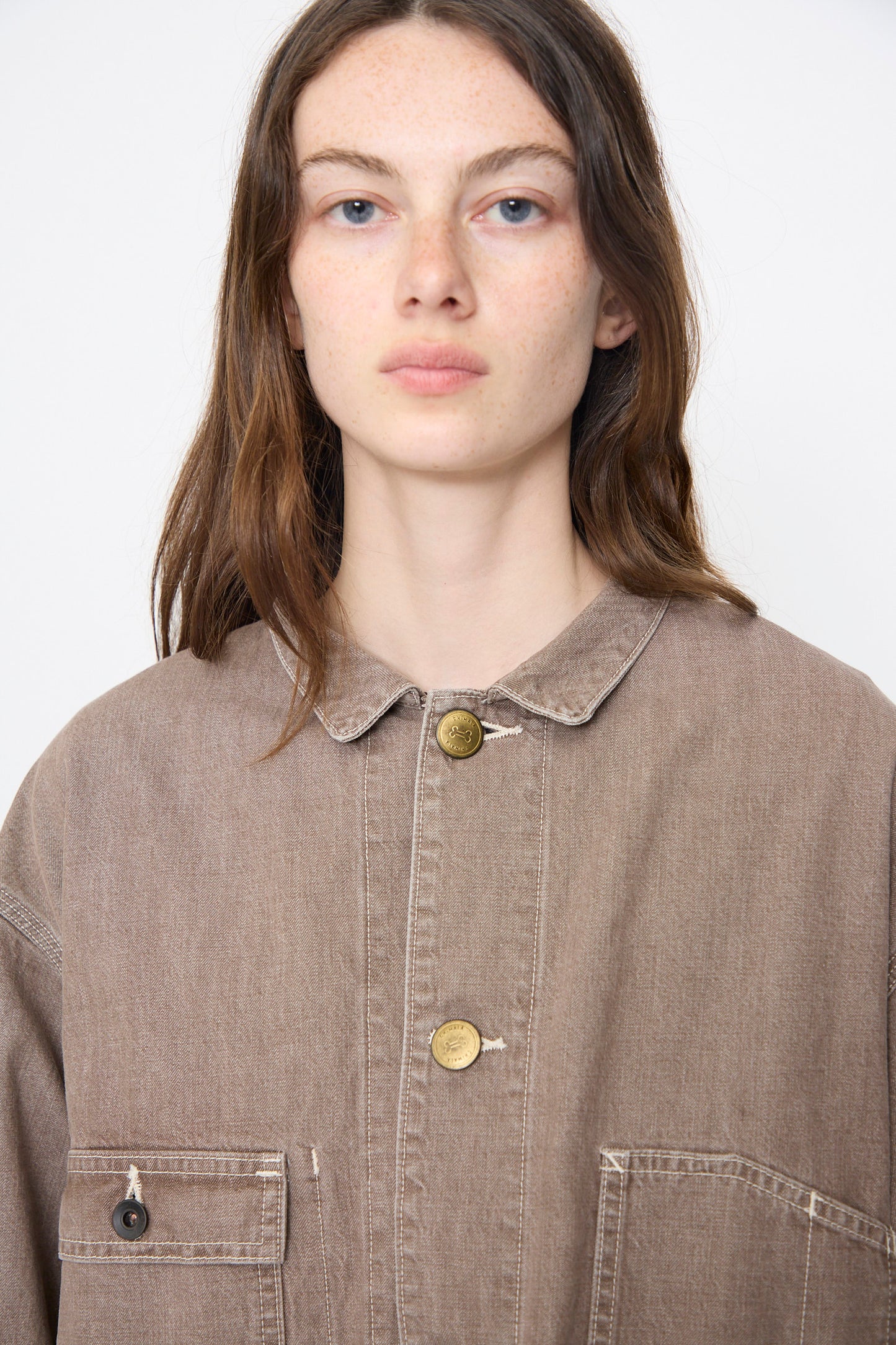 A person with long brown hair wears the Chimala Denim Short Chore Jacket in Brown and looks directly at the camera against a plain background.