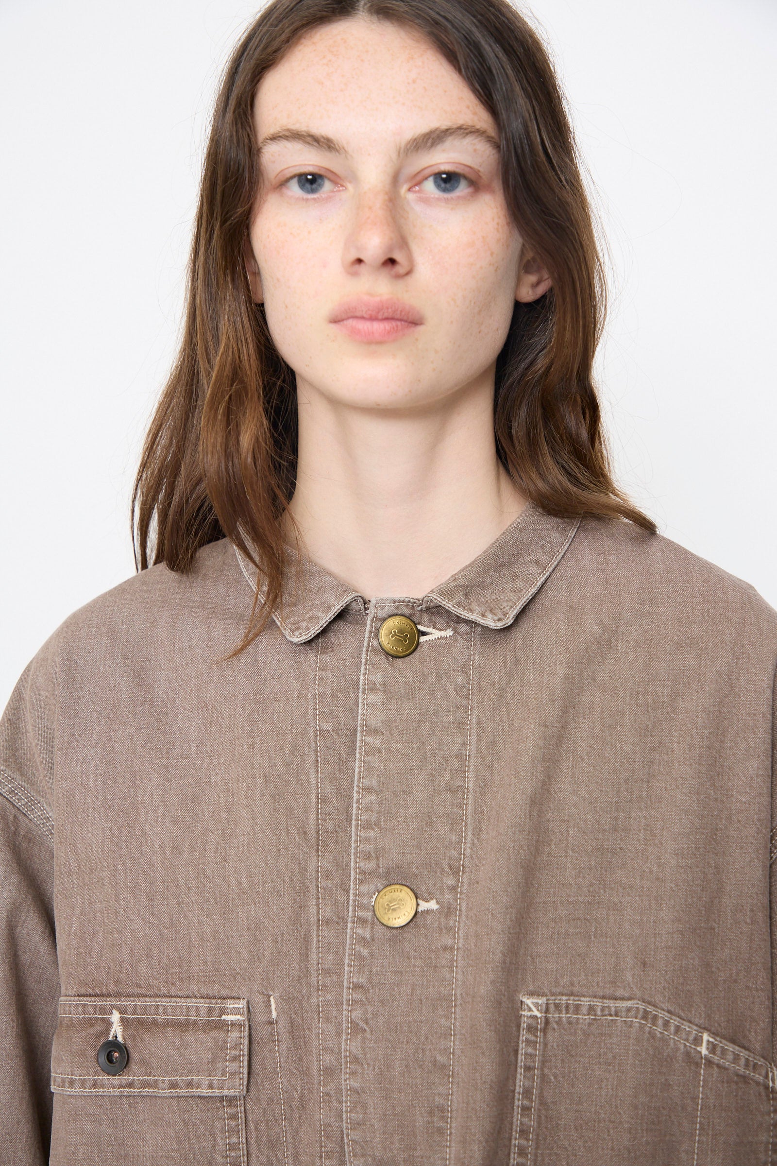 A person with long brown hair wears the Chimala Denim Short Chore Jacket in Brown and looks directly at the camera against a plain background.