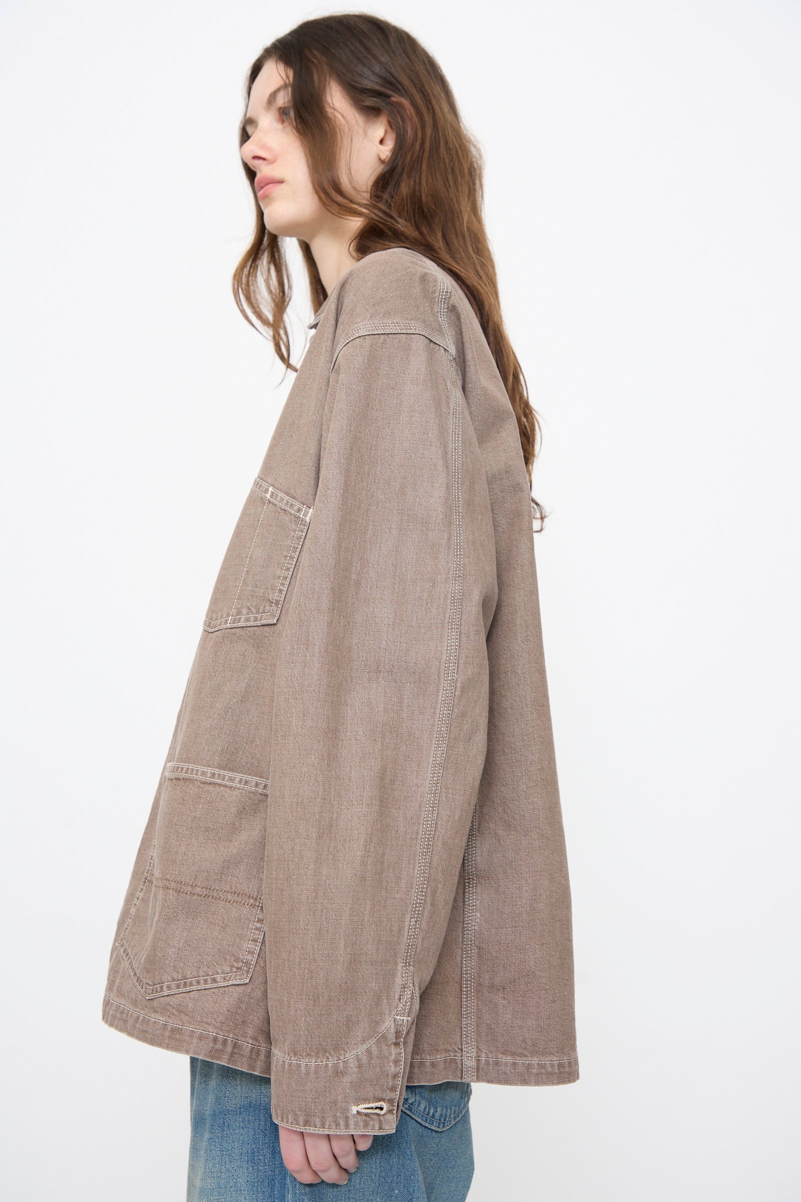 A person with long brown hair wears the Chimala Denim Short Chore Jacket in Brown—100% cotton Japanese denim with large front pockets—paired with blue jeans, standing against a plain white background.