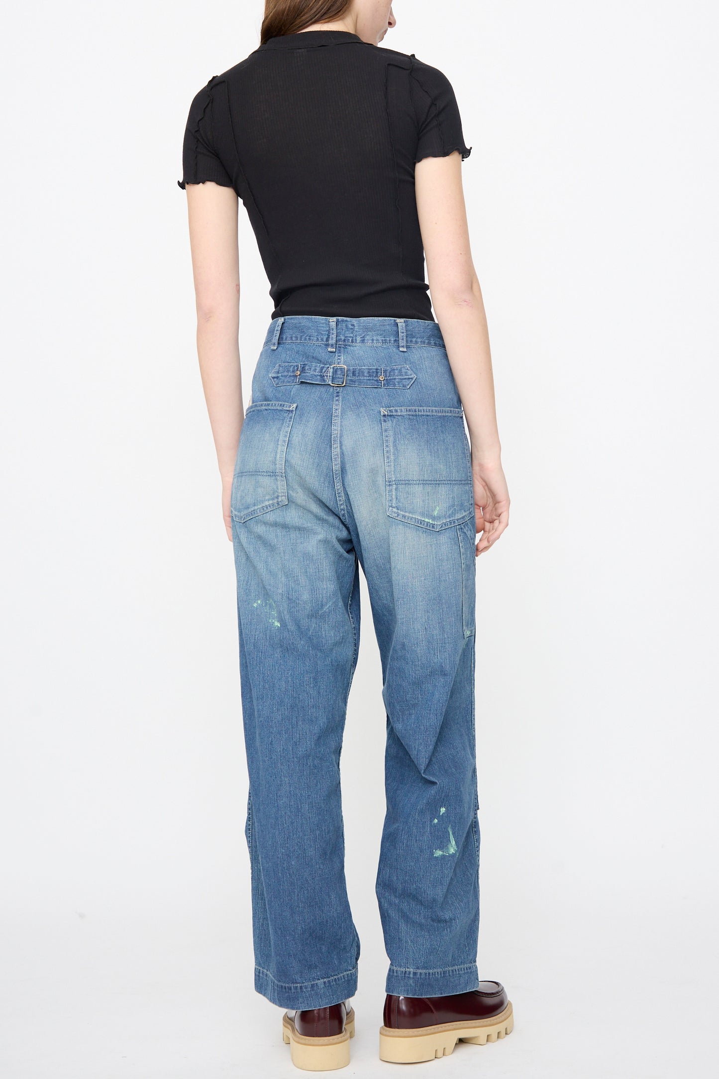 A woman stands with her back to the camera, wearing a black short-sleeve top, Chimala Denim Double Knee Painter Pants in Splushed Medium with paint marks, and beige platform shoes.