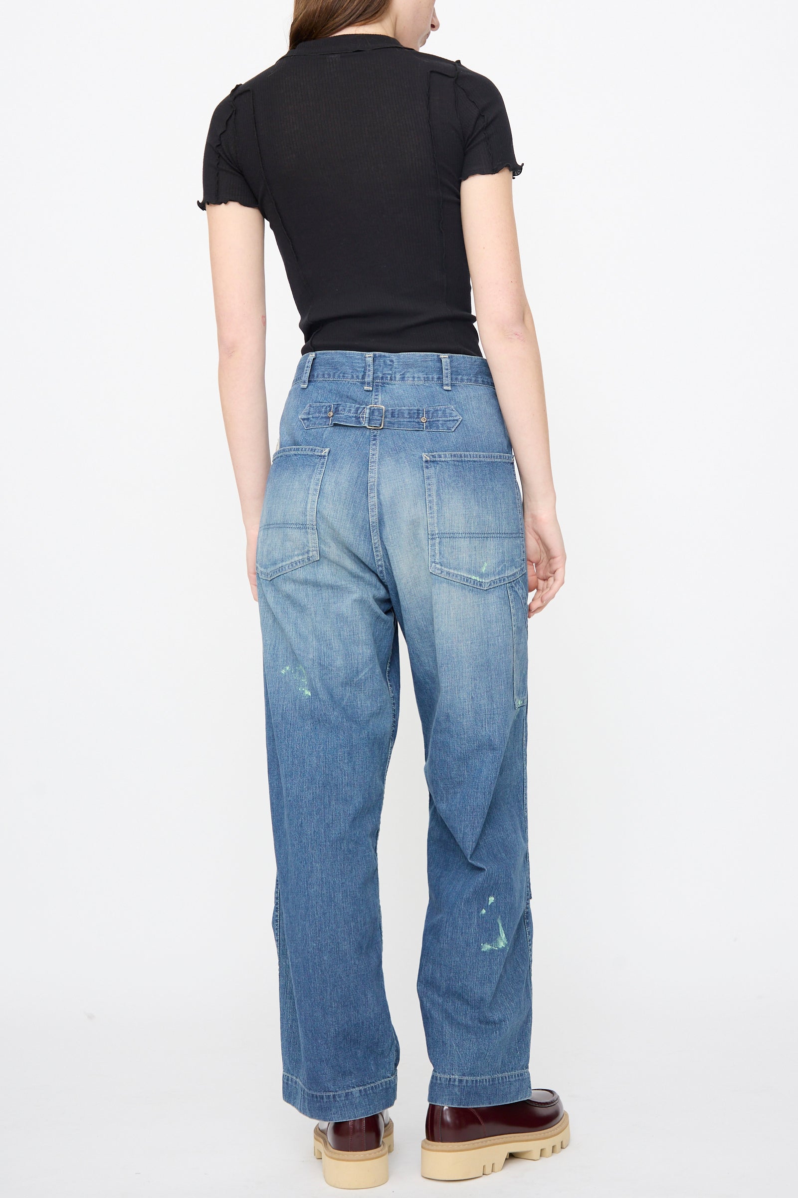 A woman stands with her back to the camera, wearing a black short-sleeve top, Chimala Denim Double Knee Painter Pants in Splushed Medium with paint marks, and beige platform shoes.