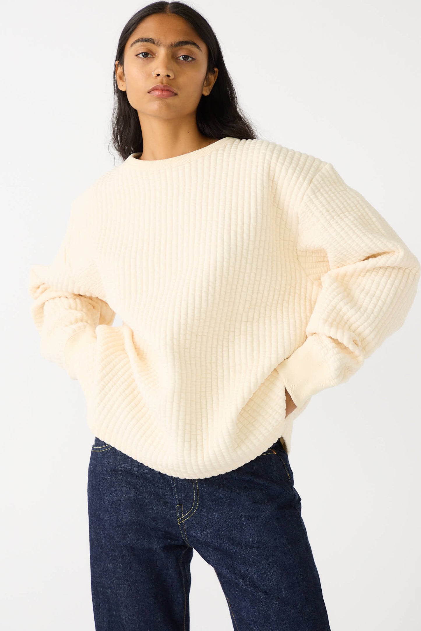 A person with long dark hair wearing Chimala's Quilted Crew Neck Top in Ivory stands with hands in pockets. This relaxed fit ensemble is completed by dark blue jeans, set against a plain white background.