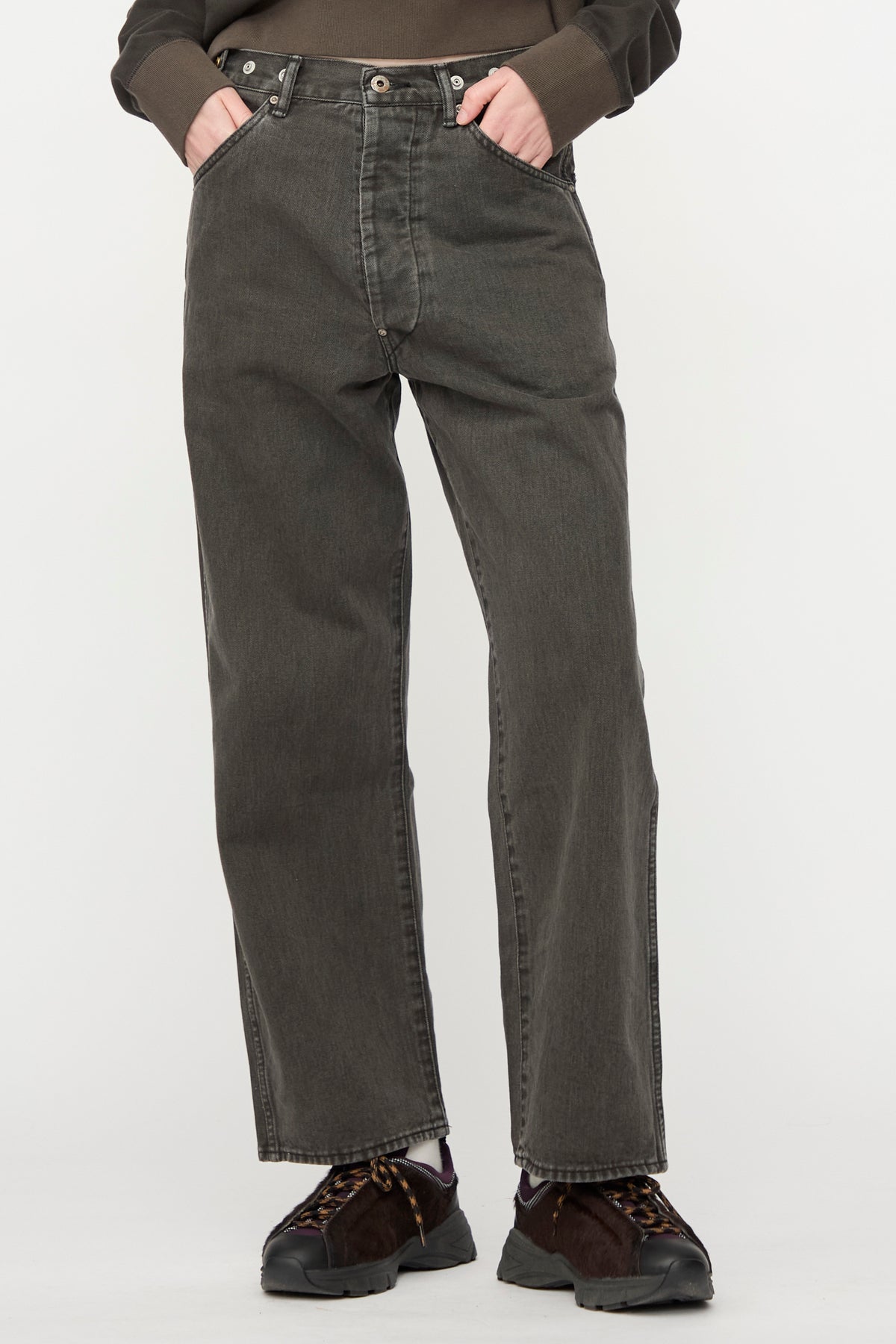 A person wearing Chimala Rope Dyed Twill Adjustable Pant in Charcoal—loose-fitting, wide-leg jeans—with hands in pockets, paired with a brown sweatshirt and dark sneakers; standing against a plain white background.