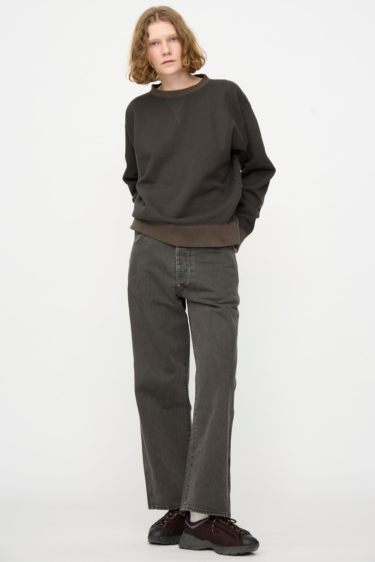 A person with shoulder-length hair stands against a plain white background, wearing a dark sweatshirt, Chimala Rope Dyed Twill Adjustable Pant in Charcoal, and dark sneakers, with one hand in their pocket.