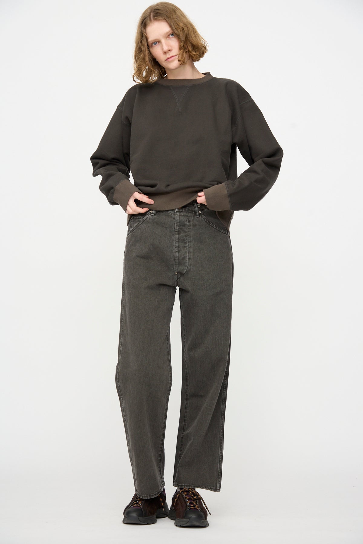 A person stands against a white background, wearing a loose dark brown sweatshirt, Chimala’s Rope Dyed Twill Adjustable Pant in Charcoal—a wide leg denim style made in Japan—and black shoes.