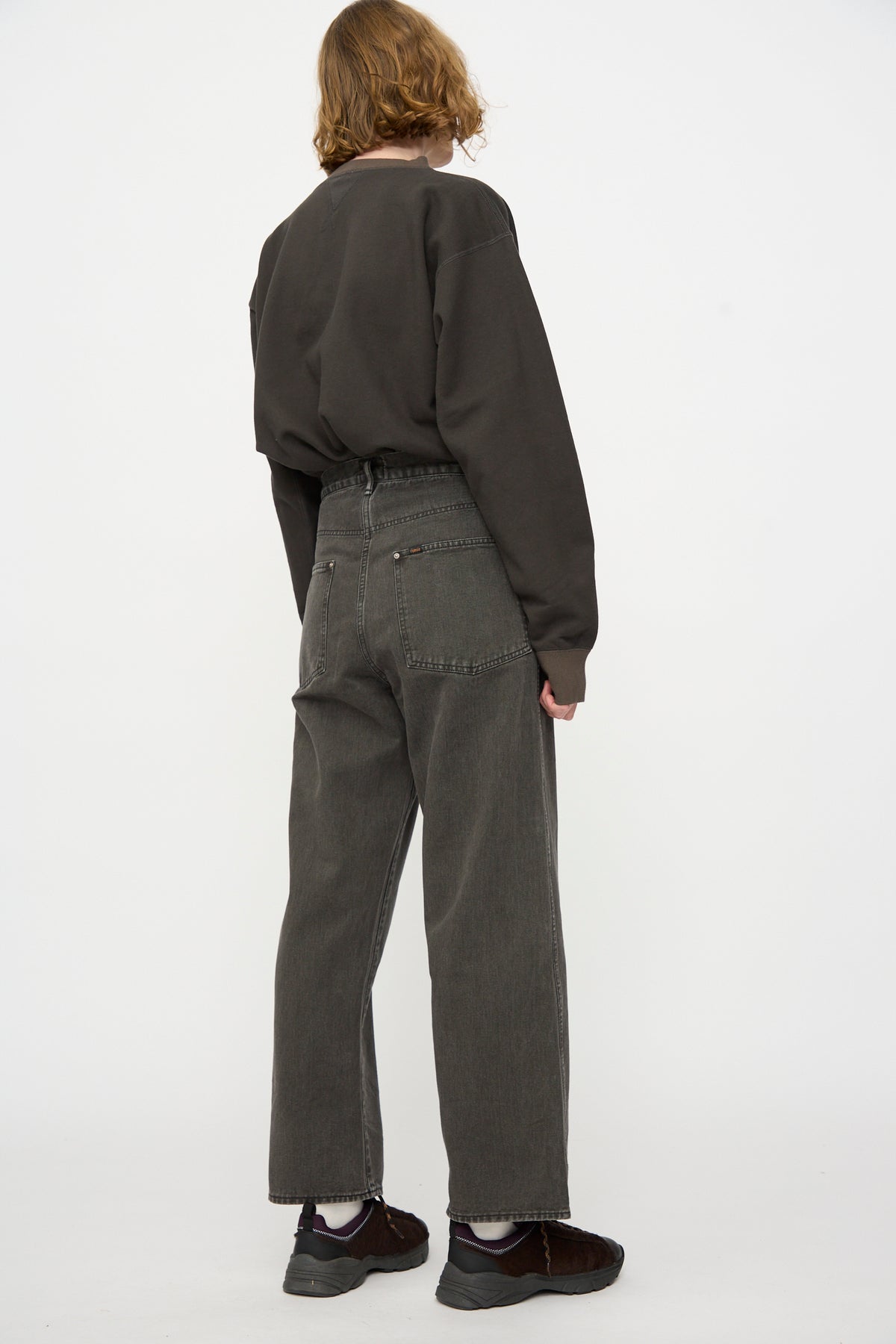 A person with shoulder-length hair stands facing away, wearing a dark long-sleeve shirt, Chimala Rope Dyed Twill Adjustable Pant in Charcoal—high-waisted wide leg denim pants made in Japan—and dark shoes against a plain white background.