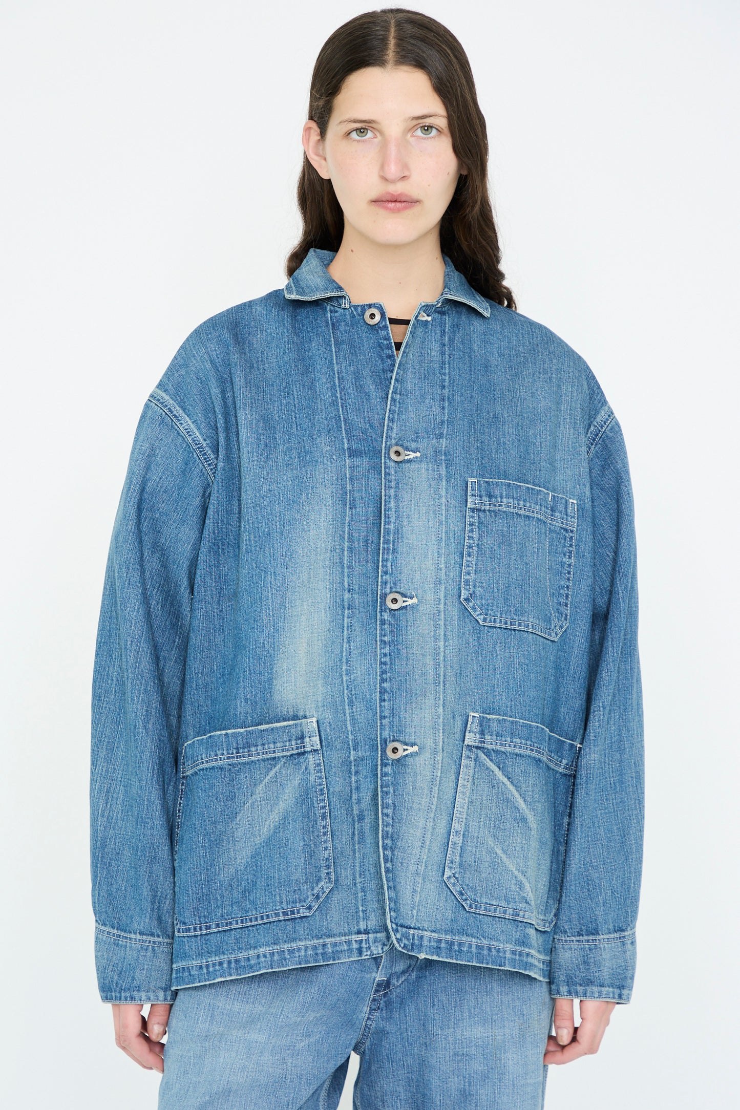 A person with long dark hair stands against a plain background, wearing the Chimala Selvedge Denim Chore Jacket in Medium Vintage.