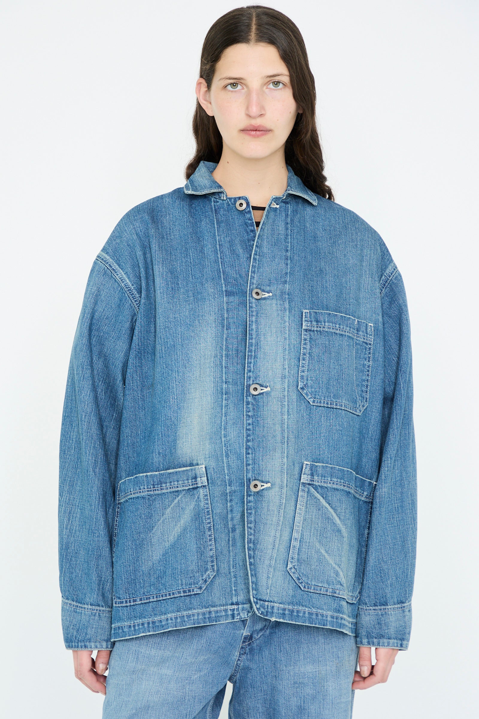 A person with long dark hair stands against a plain background, wearing the Chimala Selvedge Denim Chore Jacket in Medium Vintage.