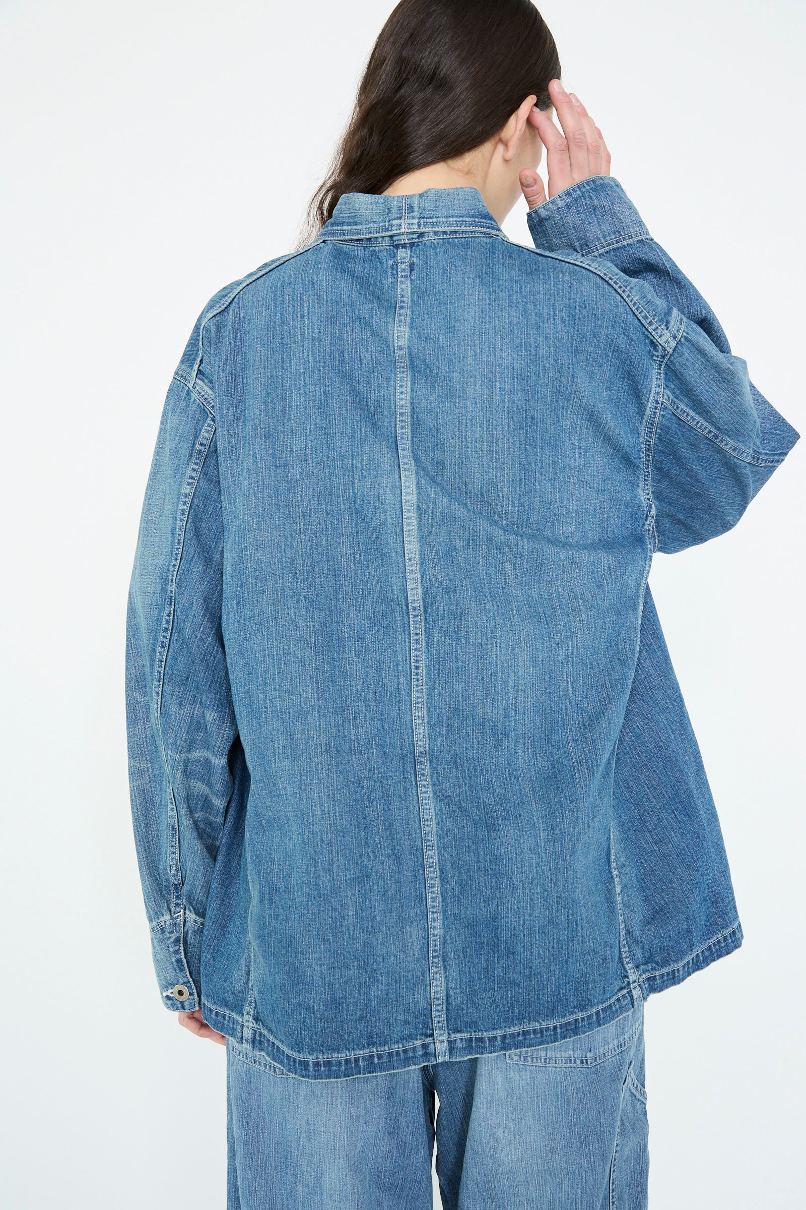 A person with long hair stands against a plain background, adjusting their hair while wearing the Chimala Selvedge Denim Chore Jacket in Medium Vintage paired with jeans.