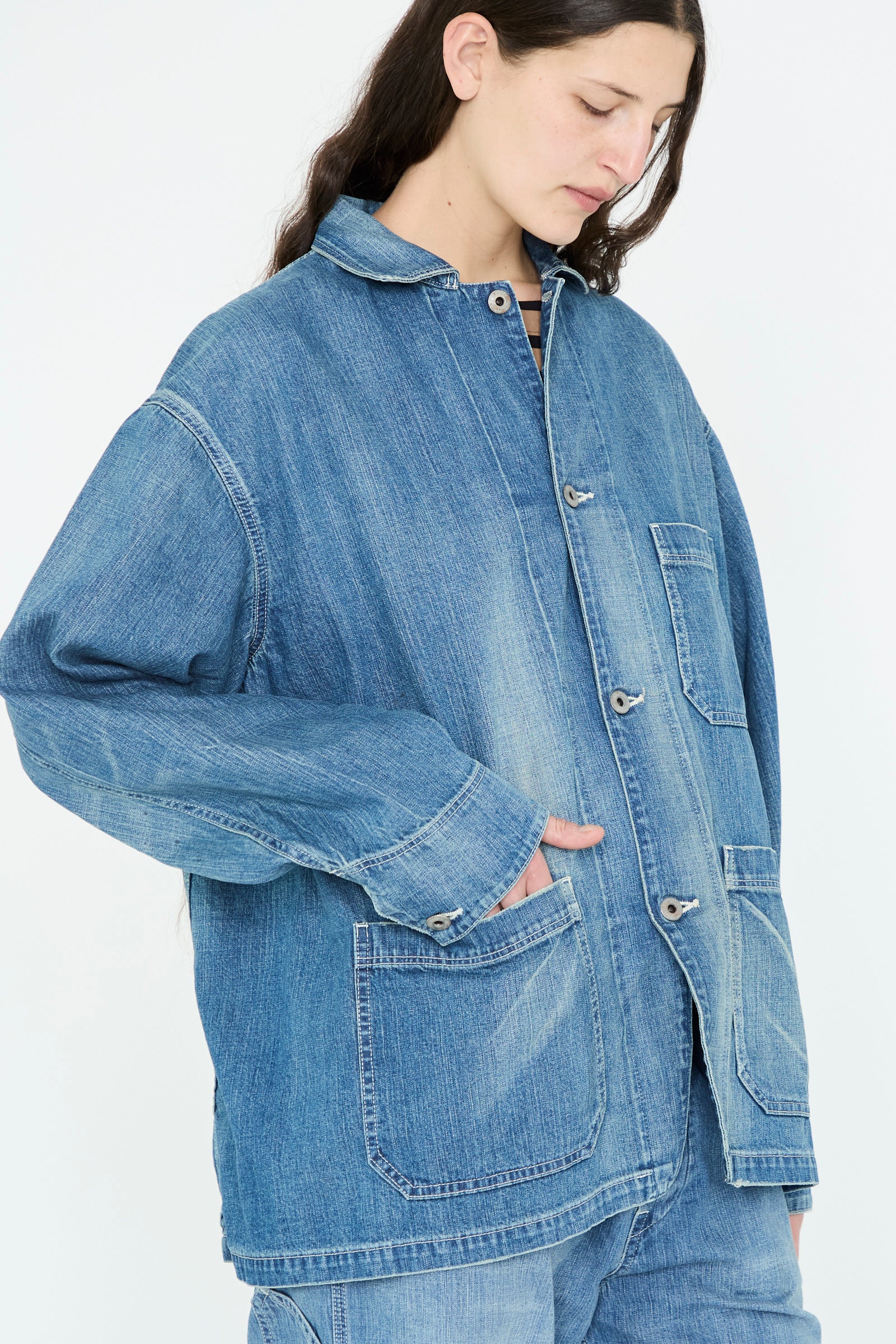 Wearing a Chimala Selvedge Denim Chore Jacket in Medium Vintage, a person looks downward against a plain background.