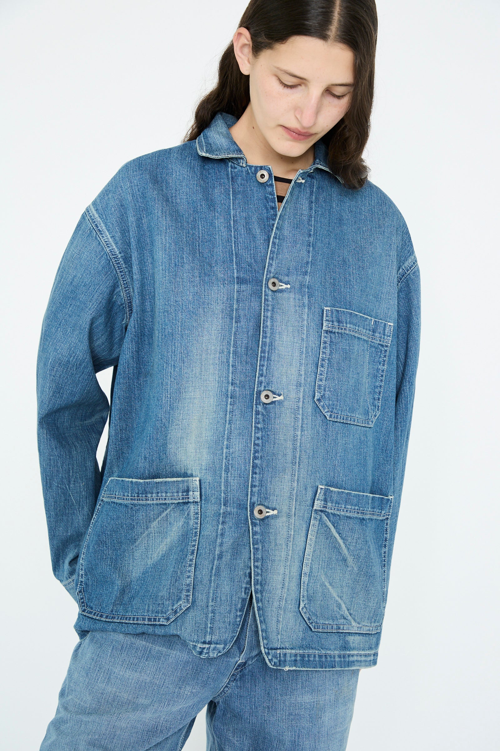Wearing Chimala's Selvedge Denim Chore Jacket in Medium Vintage, a person looks downward against a light background. The oversized jacket features button closure and large pockets, made from vintage denim.