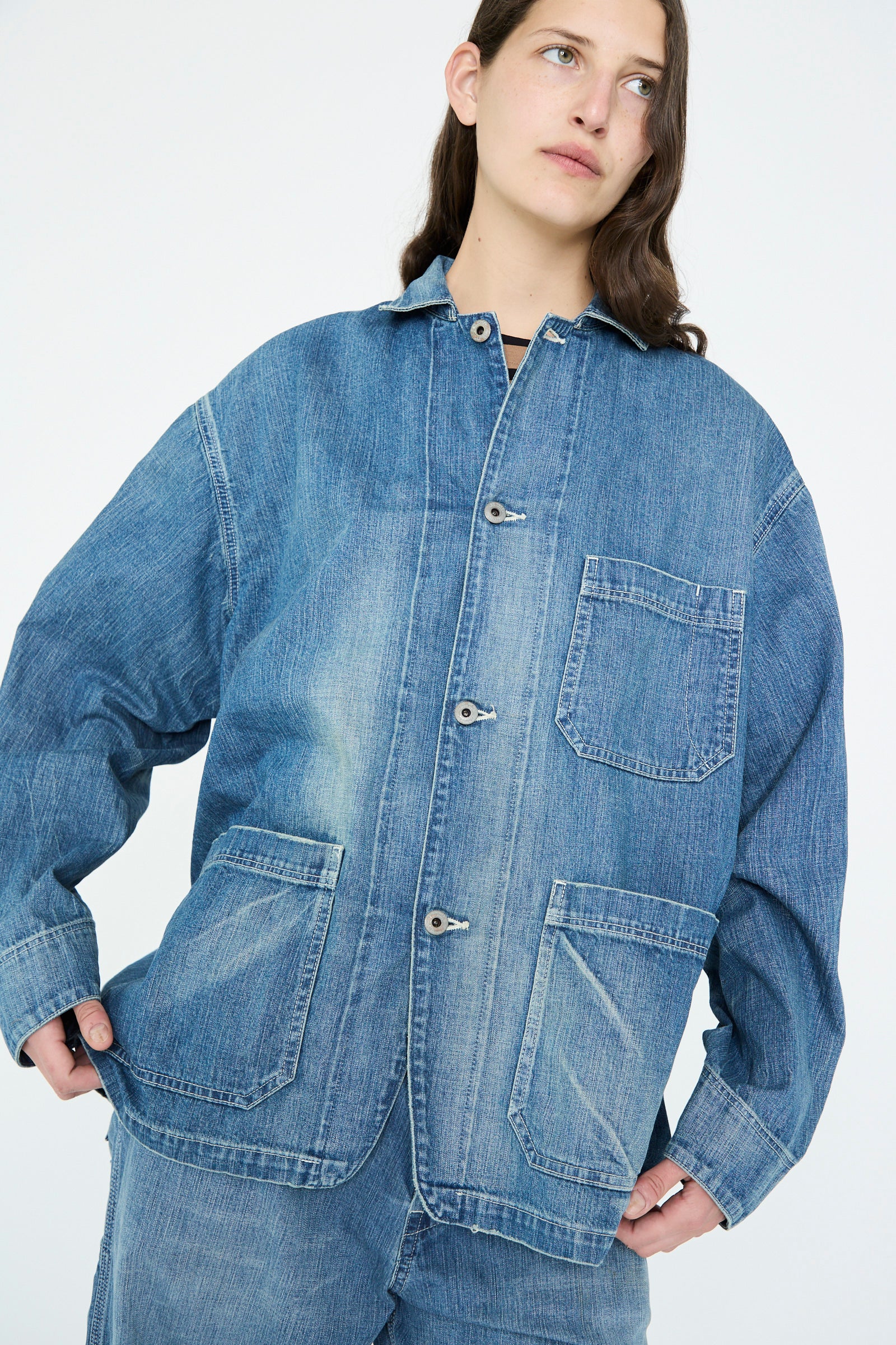 A person with long hair is wearing a Chimala Selvedge Denim Chore Jacket in Medium Vintage and matching pants, standing against a plain background.