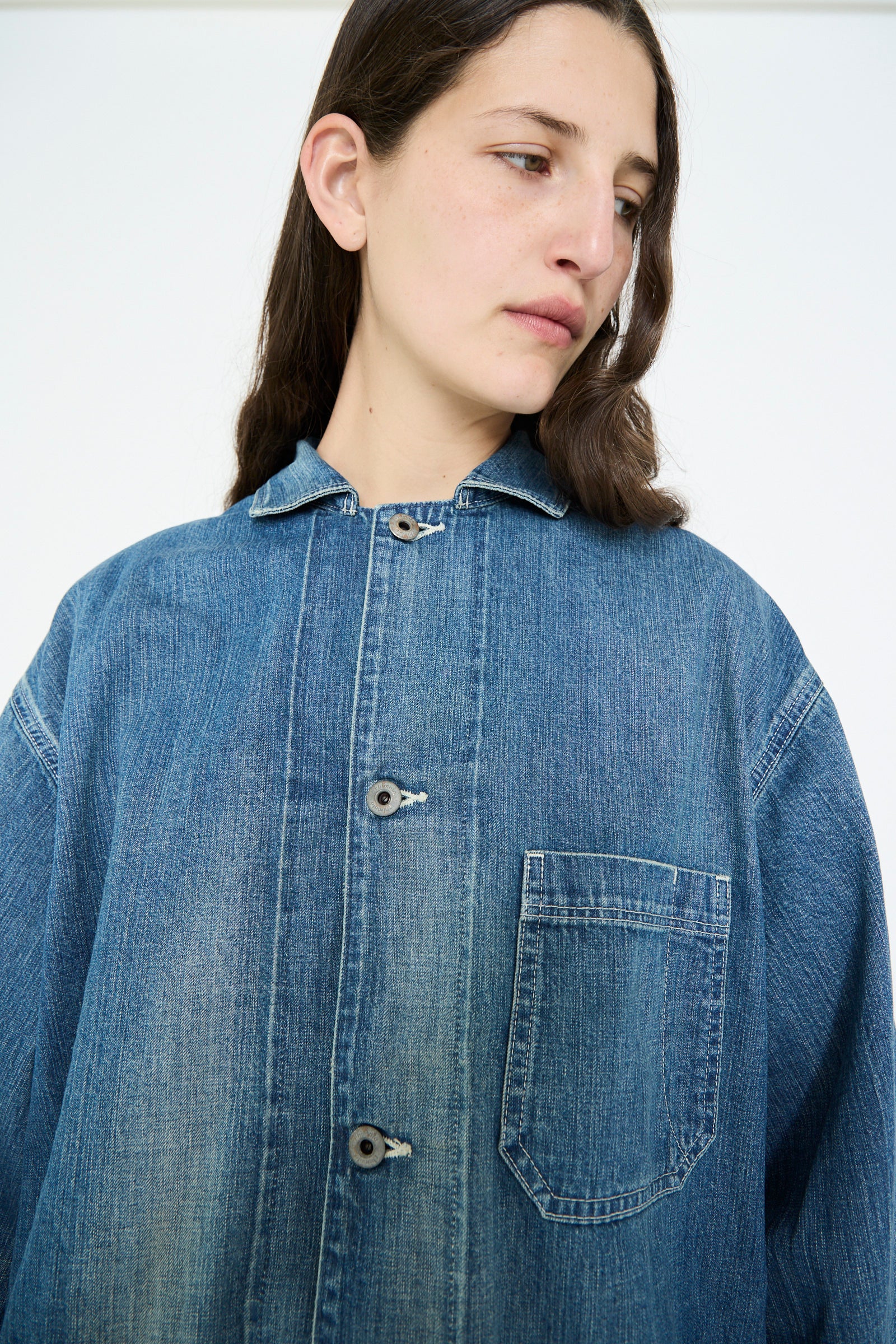 A person wears the Chimala Selvedge Denim Chore Jacket in Medium Vintage, featuring buttons and a chest pocket, gazing to the side against a plain white backdrop.