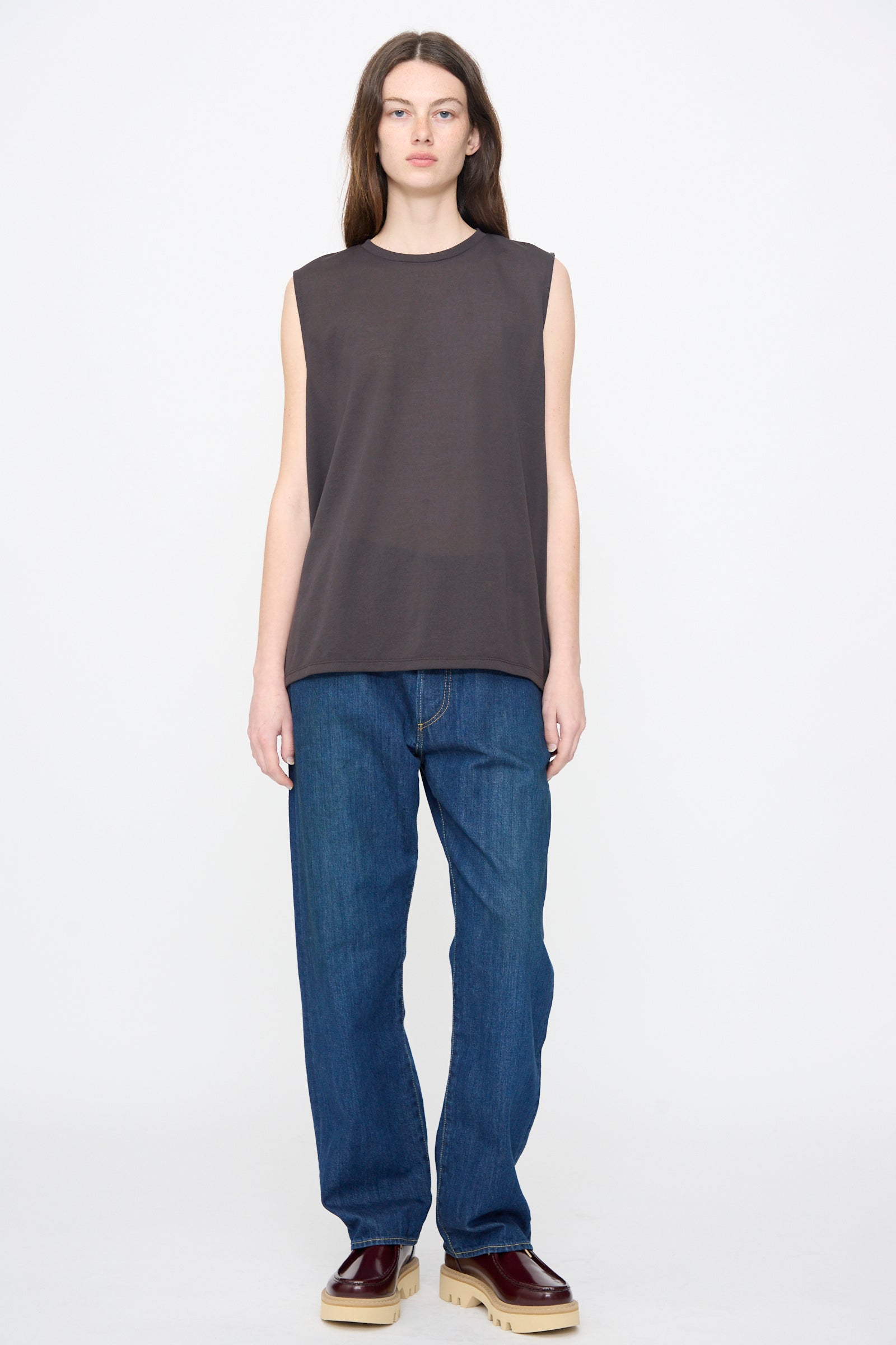 A person stands facing forward against a plain white background wearing the Chimala Sheer Fleece Muscle Tee in Charcoal, styled with blue jeans and burgundy platform shoes.