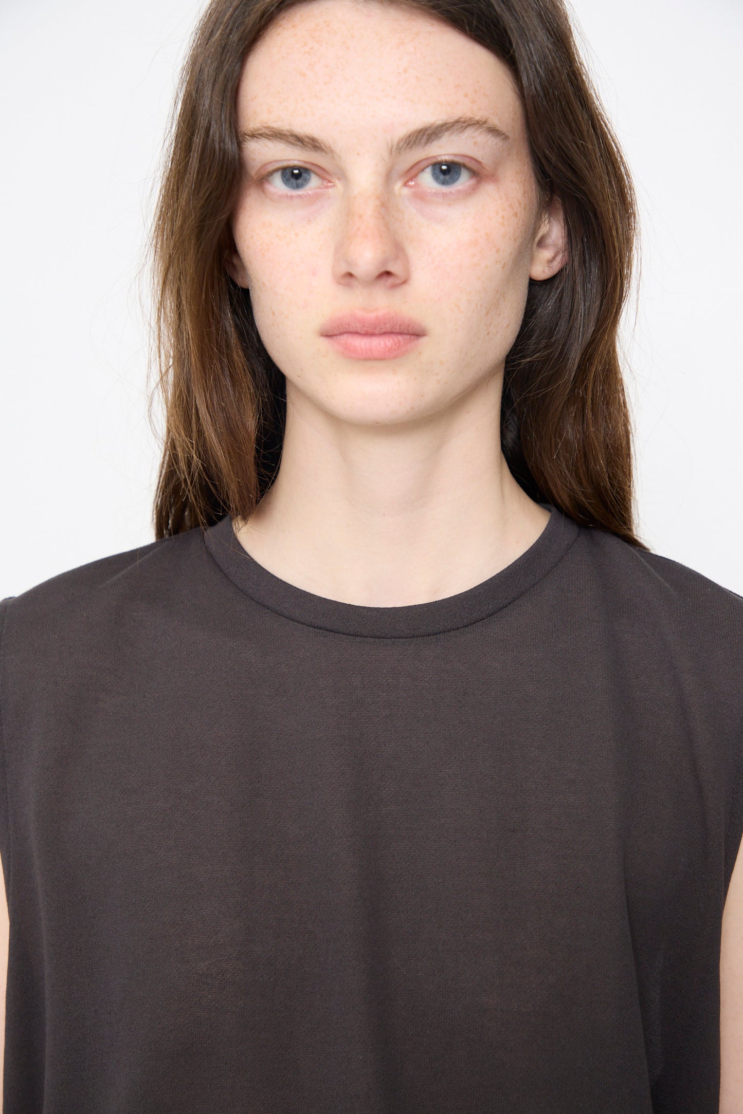 A person with long brown hair, light skin, and blue eyes wears the Chimala Sheer Fleece Muscle Tee in Charcoal, facing the camera against a plain white background.