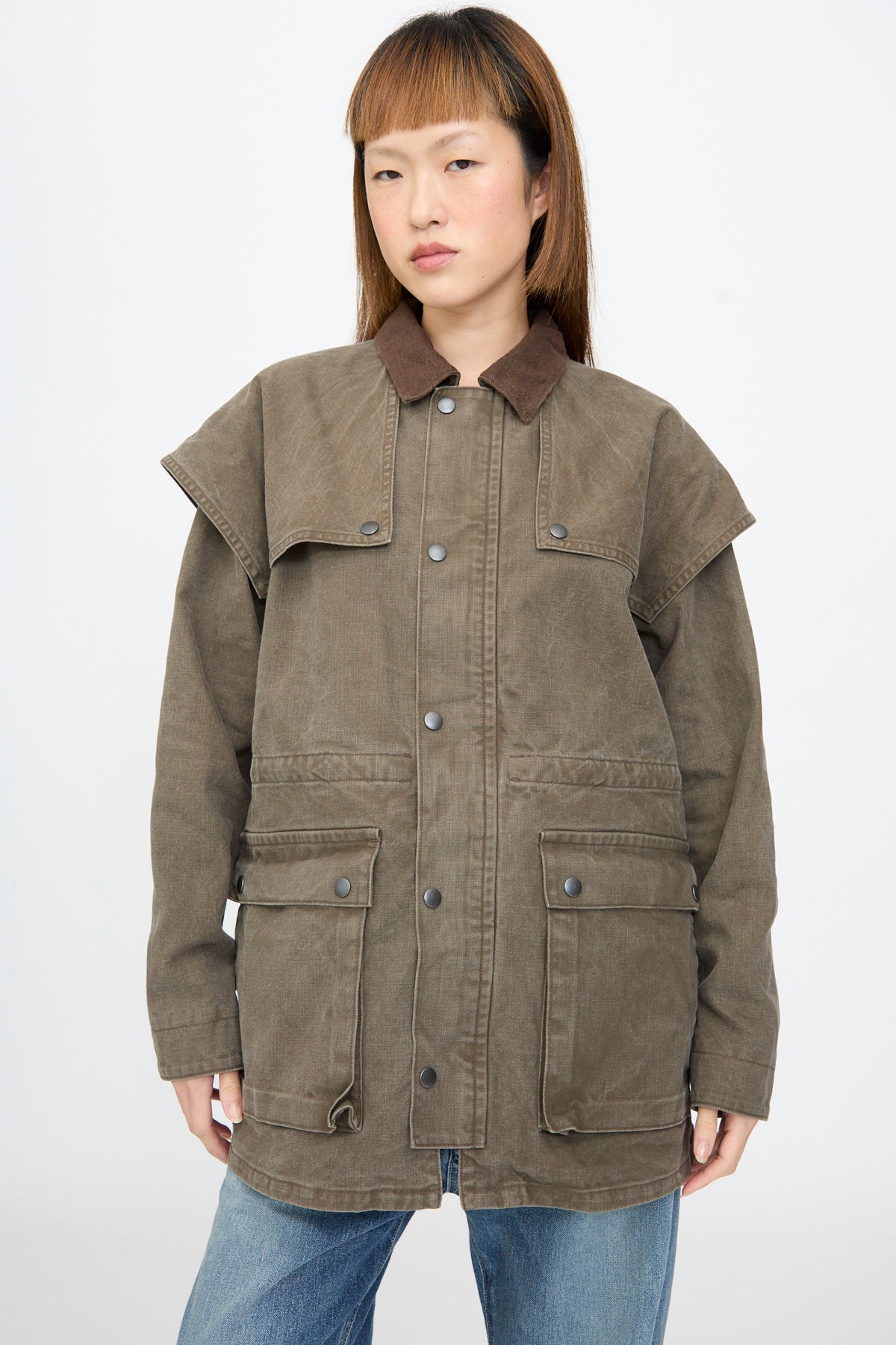 A person wearing a brown Chimala Sulfer Dyed Canvas Lumber Coat featuring a suede collar, layered shoulder panels, and multiple pockets.