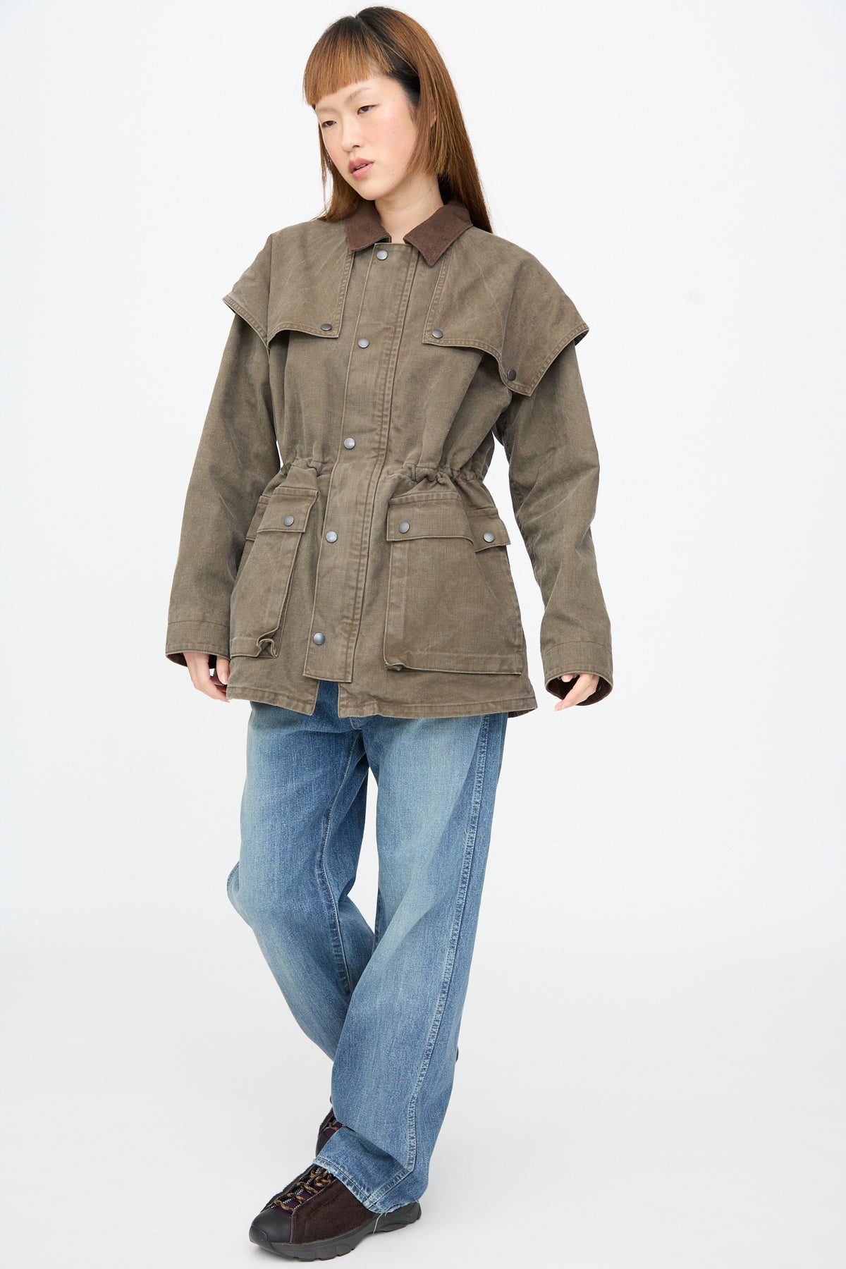 A person wears a Chimala Sulfer Dyed Canvas Lumber Coat in brown featuring a suede collar, wide shoulder panel, and drawstring waist, paired with blue jeans.