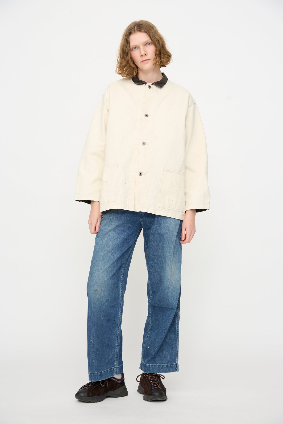 A person stands against a plain white background wearing the Chimala Twill Engineer Jacket in Ivory Splatter—made in Japan—paired with blue jeans and black shoes, looking at the camera with a neutral expression.